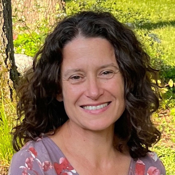 Jennifer Rubbo, a relaxed person with curly dark brown hair, wearing a floral top and sitting outdoors among trees.