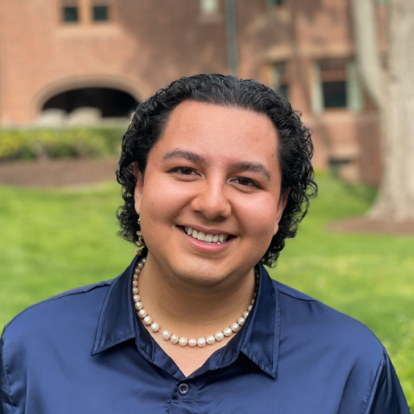 Julian Aguilar, a cheerful person with short curly hair, wearing a navy blue shirt and a pearl necklace, standing on a campus lawn with a brick building behind.