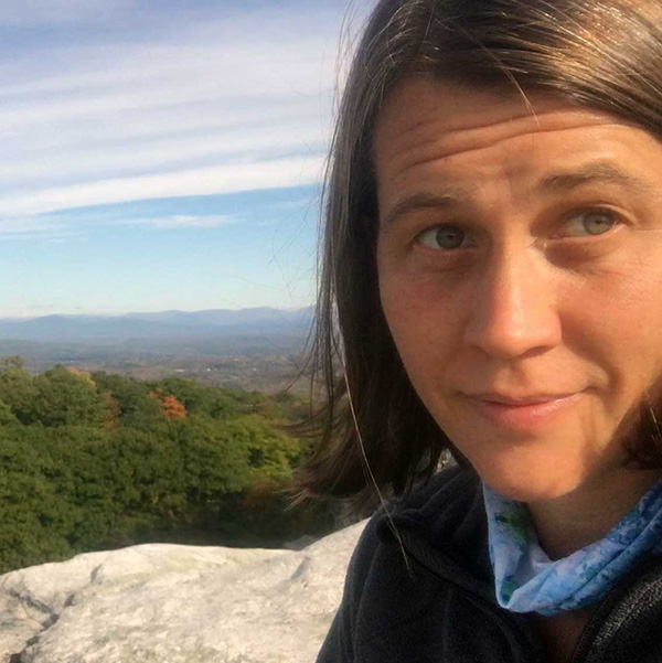 Keri VanCamp, a thoughtful-looking person with straight brown hair, wearing a fleece jacket and neck gaiter while sitting on a rock ledge with a mountain view behind.