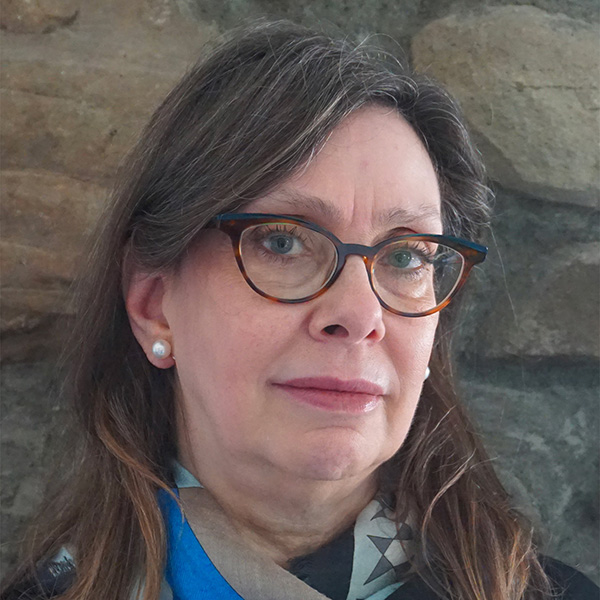 Yvonne Elet, a person with long brown hair, wearing tortoiseshell glasses and pearl earrings.