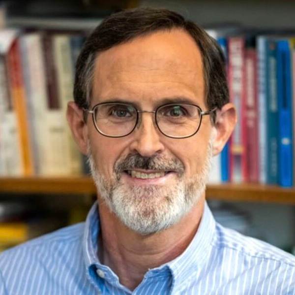 Kenneth Foster, a smiling person with short dark hair and a gray beard, wearing glasses and a blue striped shirt, in front of a blurred background of bookshelves.