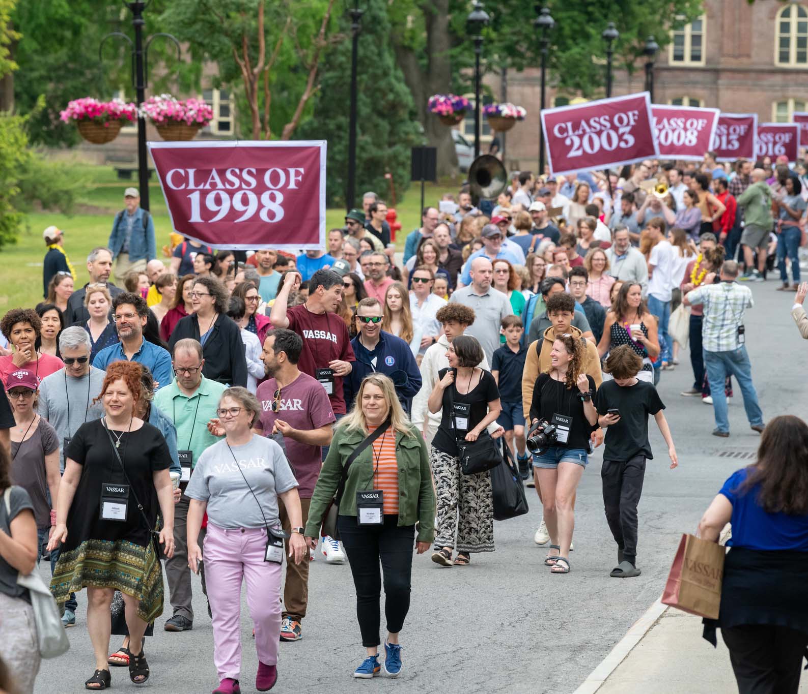 A large group of people walking along a campus road, with some banners displaying class years.