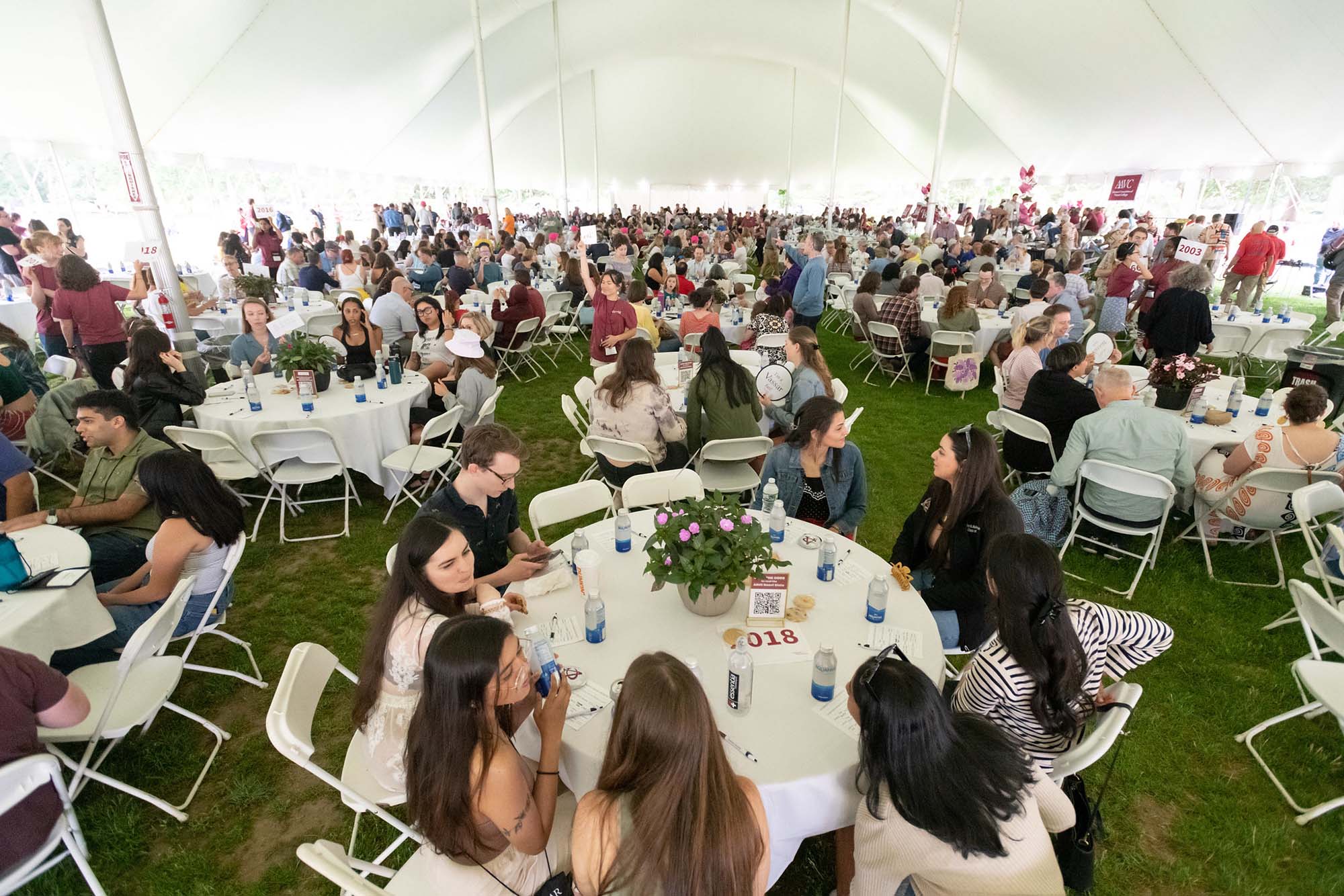 A large group of people sitting at round tables, coversing with one and other, under a huge white tent.