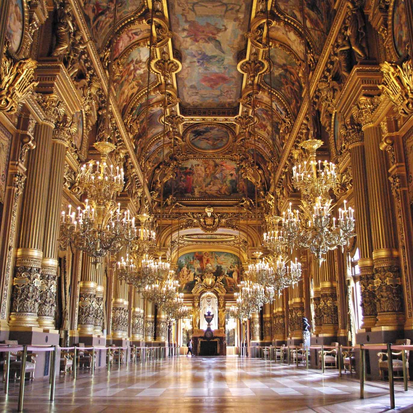 Palais Garnier in Paris, a lavish hall adorned with gold trim, ornate chandeliers, and ceiling frescoes, with symmetrical columns and mirrors lining the sides beneath a vaulted painted ceiling.
