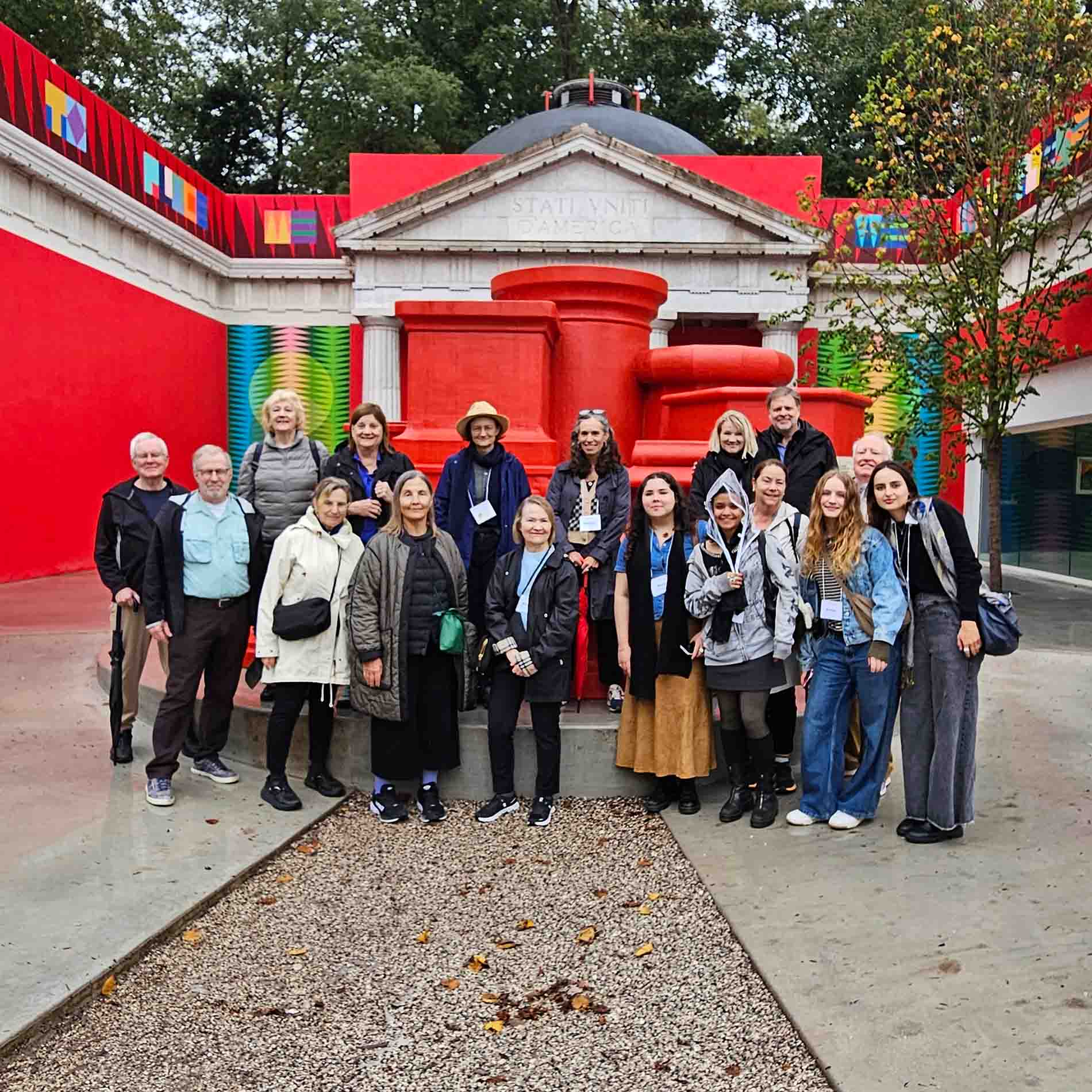 A group of people pose together in front of a vividly colored, modern art pavilion featuring bold red sculptural elements and a classical-style pediment inscribed with 