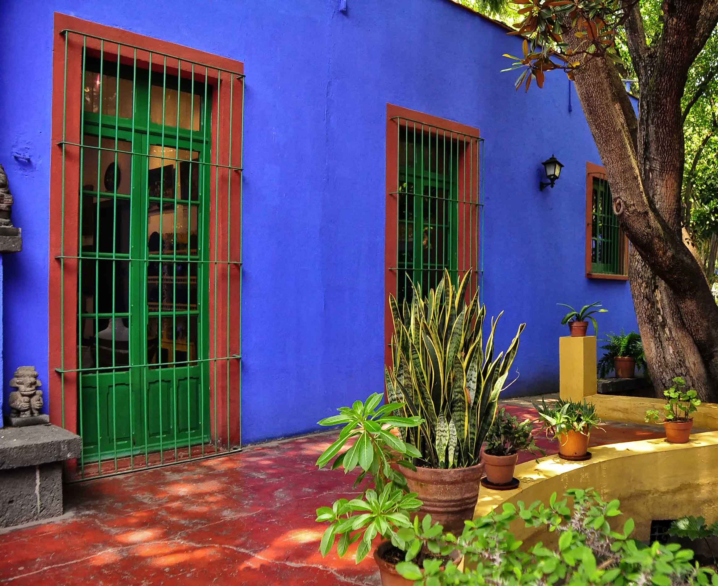 Frida Kahlo House in Mexico City, a vibrant cobalt blue exterior wall with green-paned windows framed in red, surrounded by lush potted plants and shaded by trees in a courtyard with red tile flooring.
