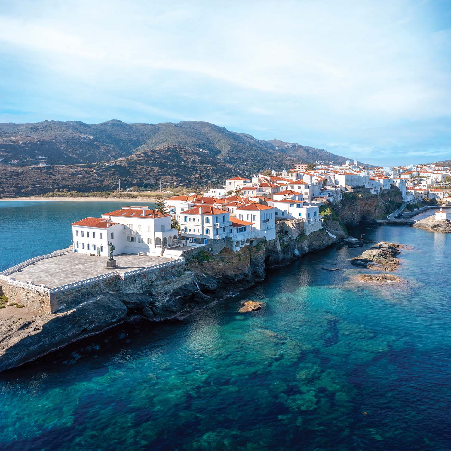 Chora in Andros, Greece, a coastal village of whitewashed buildings with terracotta roofs perched on rocky cliffs above crystal-clear blue water, backed by rolling hills under a bright sky.