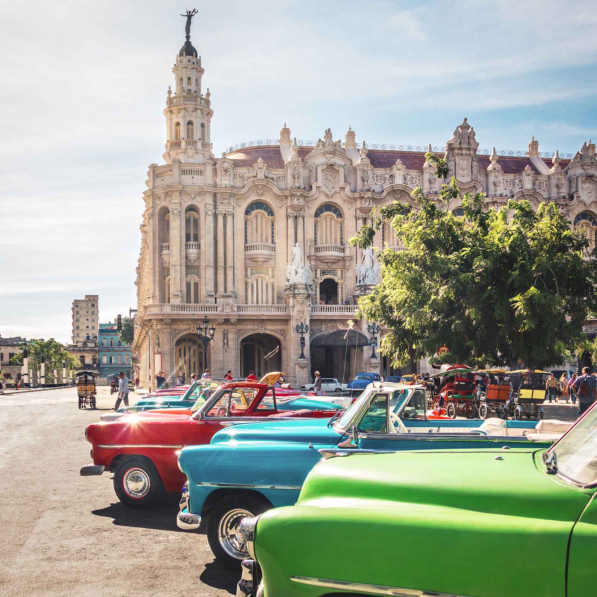 A row of colorful vintage convertible cars parked on one side of a road in front of an elaborately decorated multi-story building on a sunny day.