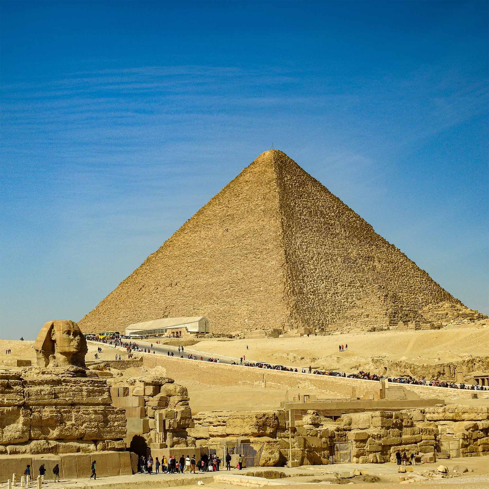 A very large stone pyramid is on a hill above a stone sphinx and stone walls of varying heights, on a sunny day.