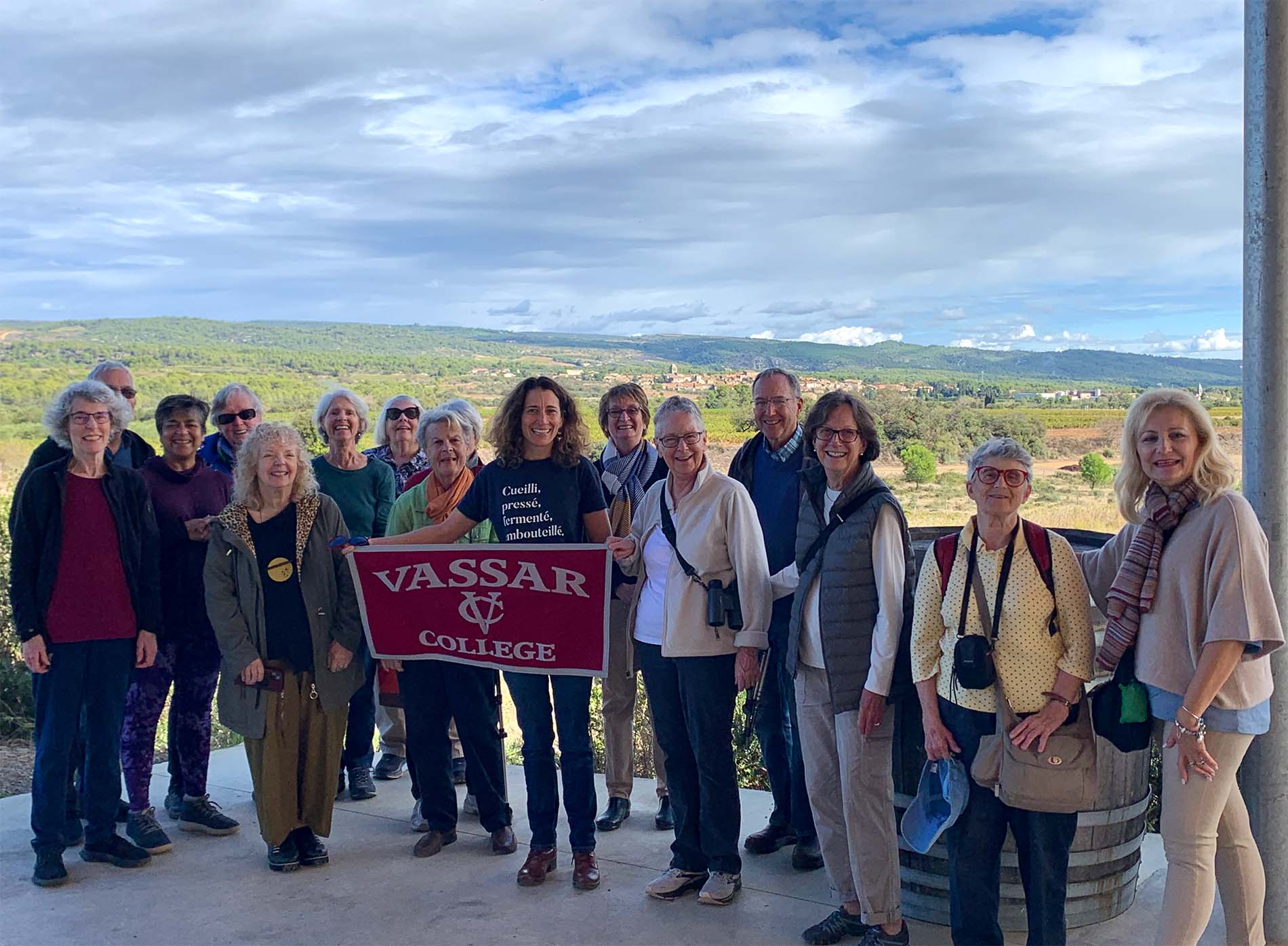A group of people, one holding a Vassar College banner, standing and smiling at the viewer with a sunny green countryside and cloudy skies behind them.