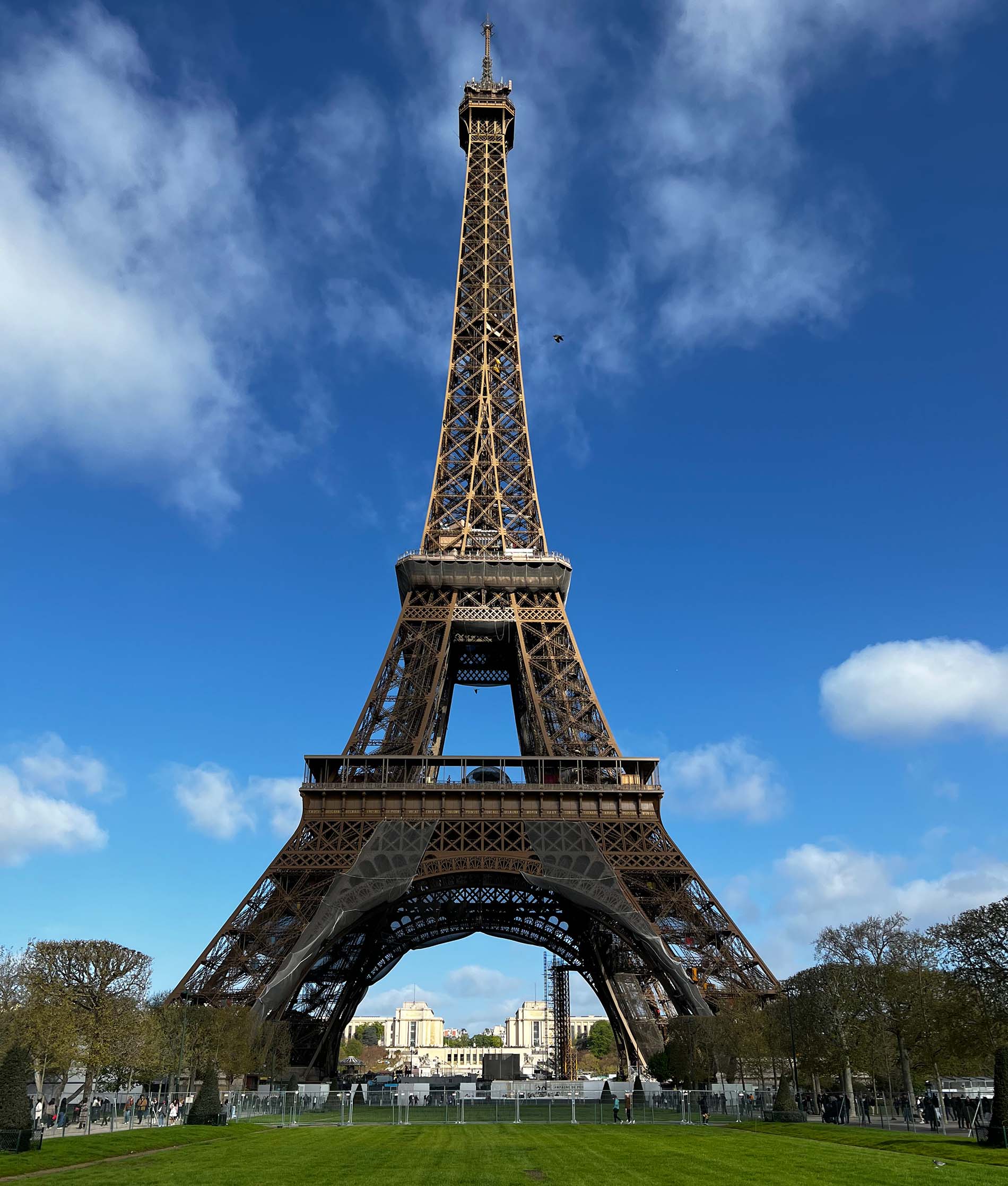 The Eiffel Tower, a tall open metalwork structure with sloping sides narrowing to a spire on top, with platforms on several levels, standing at the end of a large green field. Photo Credit Susan Brkich ’86.