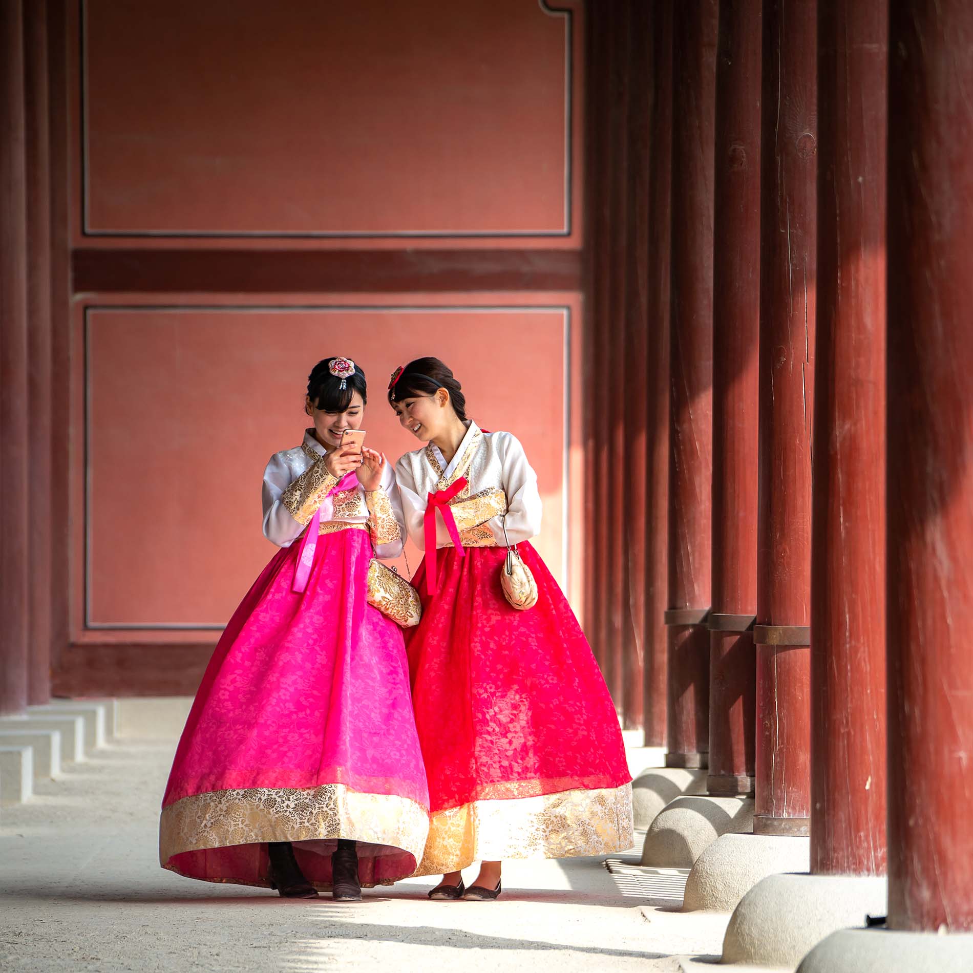 Two people wearing long gowns with gold collars, cuffs and trim and pink or red skirts stand looking at a cell phone and smiling on a wide path between two rows of tall red wooden pillars with a red wall behind them.