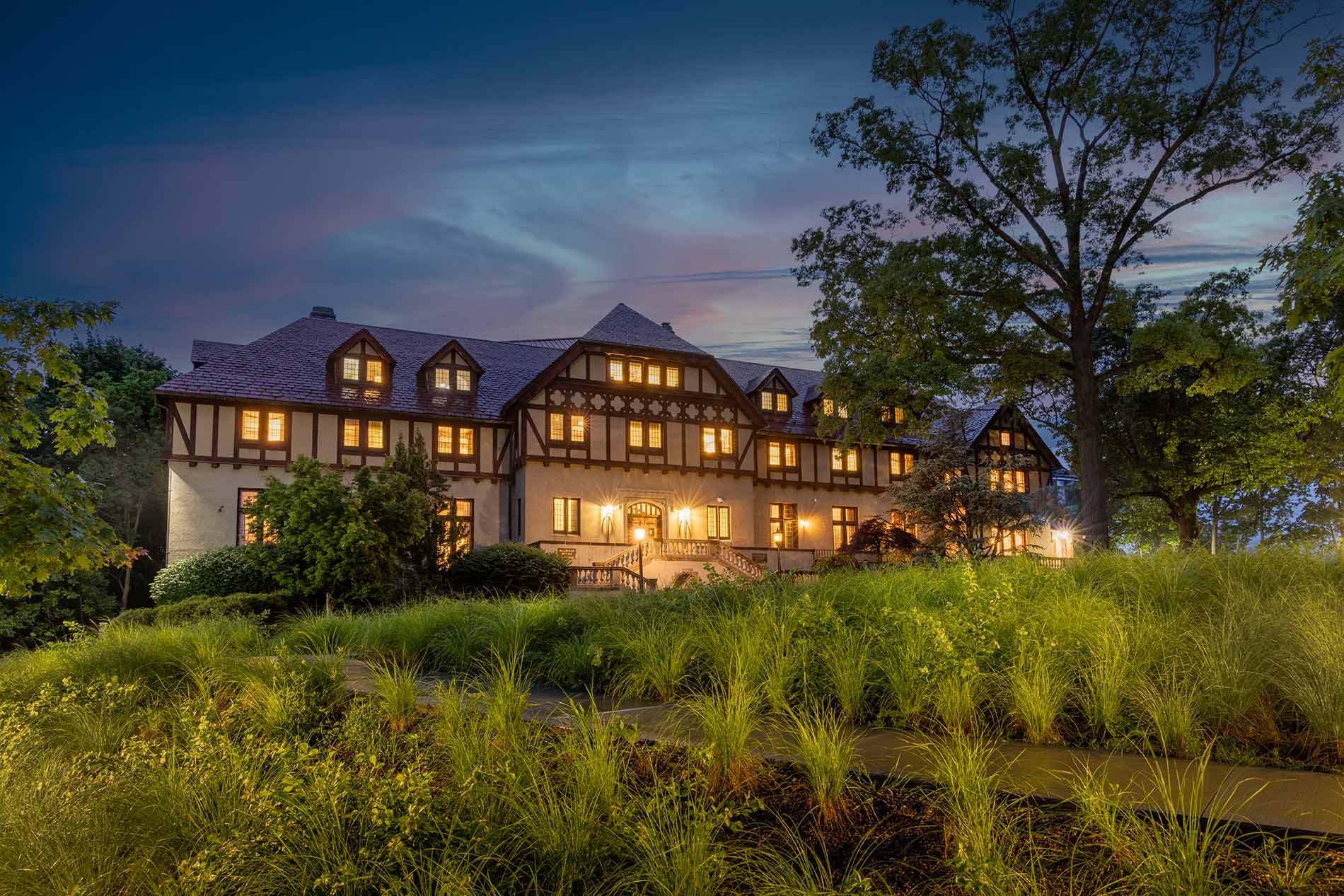 A three-story tudor-style building on a hill lit warmly at dusk, surrounded by tall trees and lush landscaping.