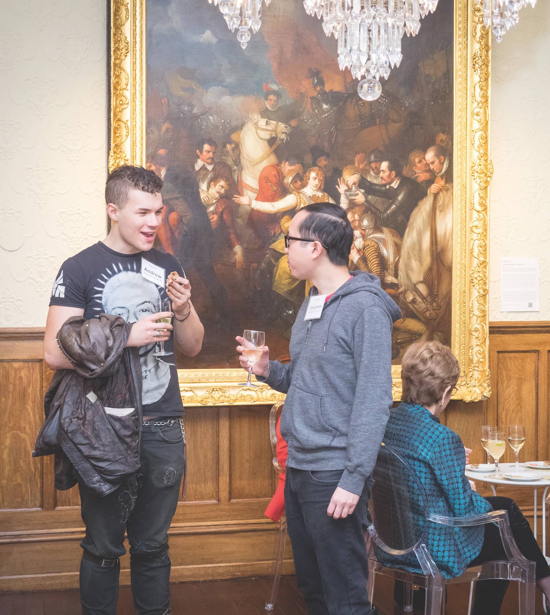 A person in a black shirt and black pants stands next to a person in a grey sweater and black pants as they converse in front a large painting.