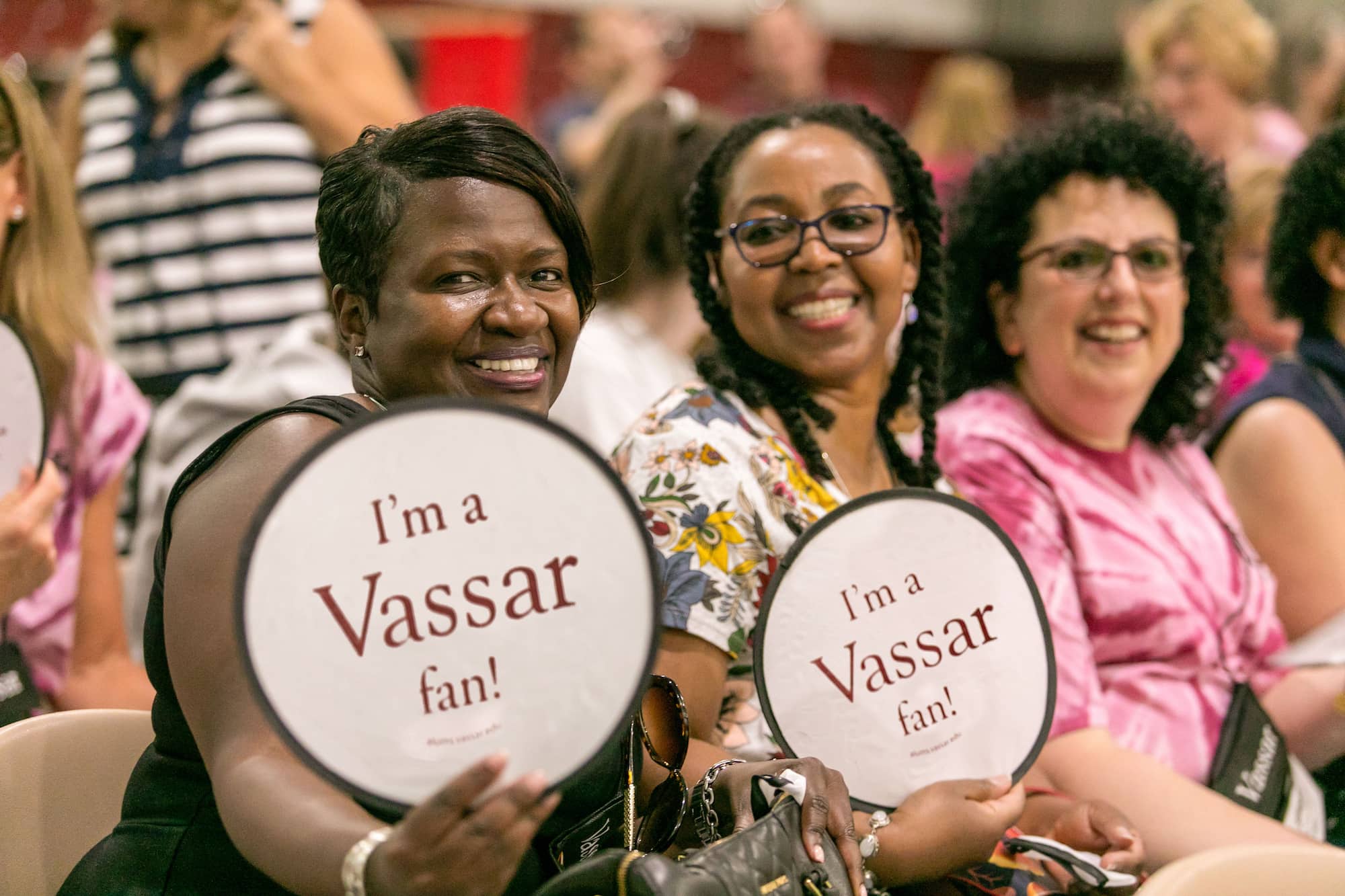Three smiling people in an audience. Two are holding I’m a Vassar Fan! signs.