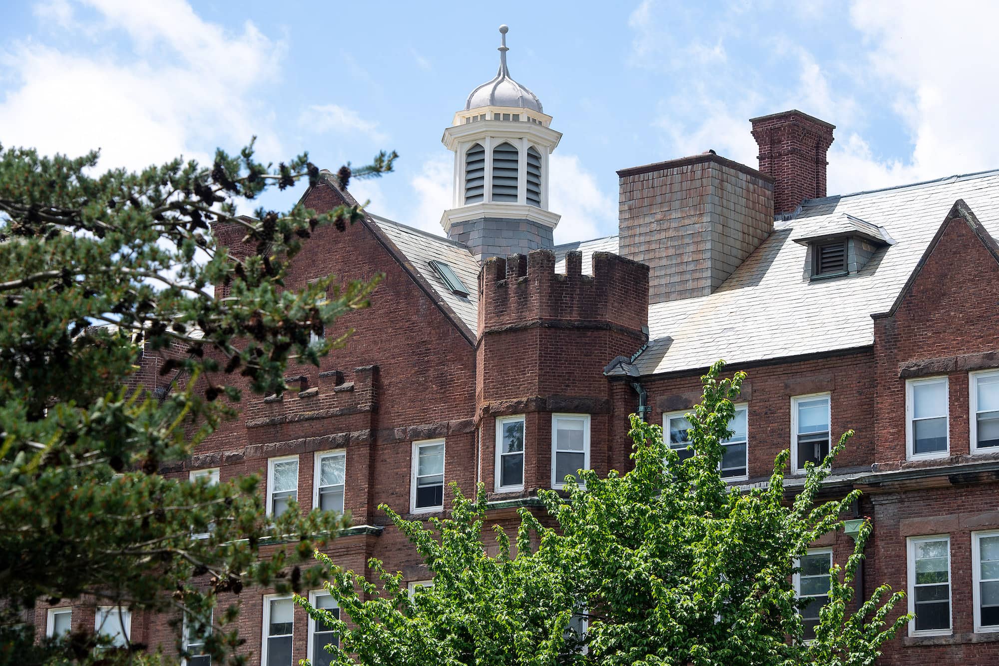 A multi-storied red brick building with a light slate roof and a round cupola, viewed through the green leaves of some trees.