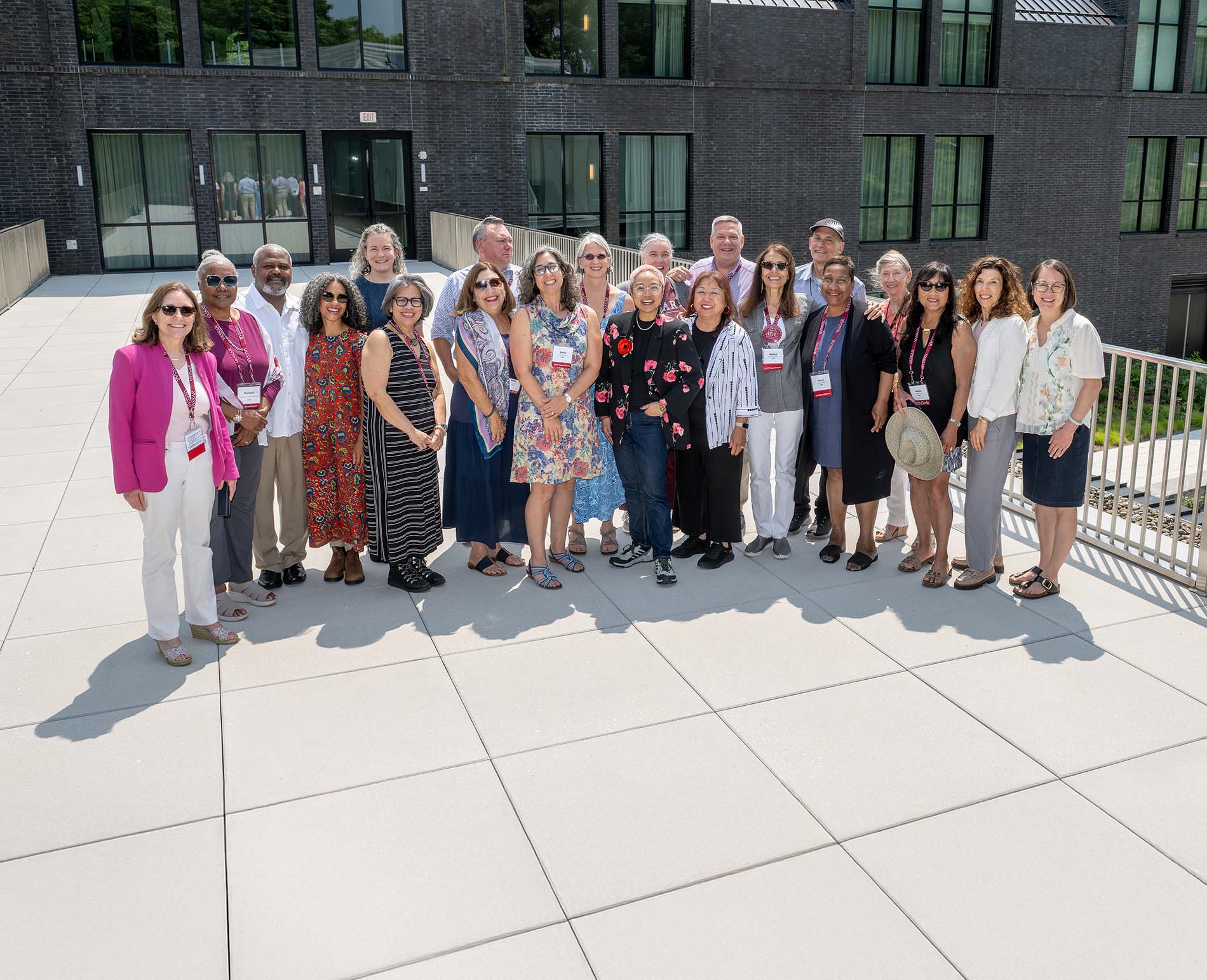 A diverse group of people stands close together outdoors, smiling at the camera in bright sunlight, wearing name badges and casual summer clothing. A modern brick building with large windows is in the background.