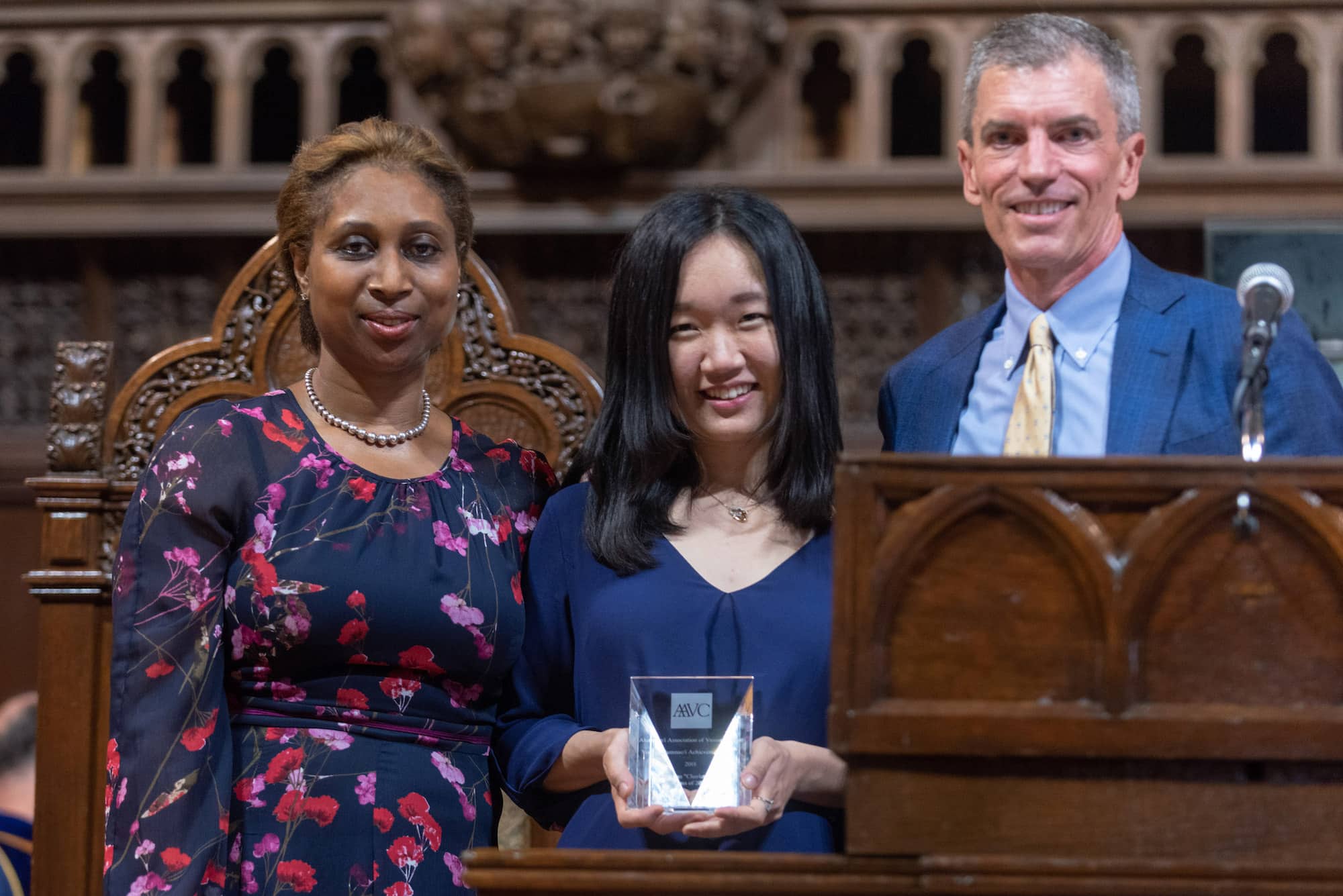 Three people stand next to each other. On the left is a person in a blue outfit with light hair, in the middle is a person with mid length dark hair in a blue shirt and on the right is a person with short white hair in a blue suit. The person in the middle is holding an award while all three people smile at the viewer.