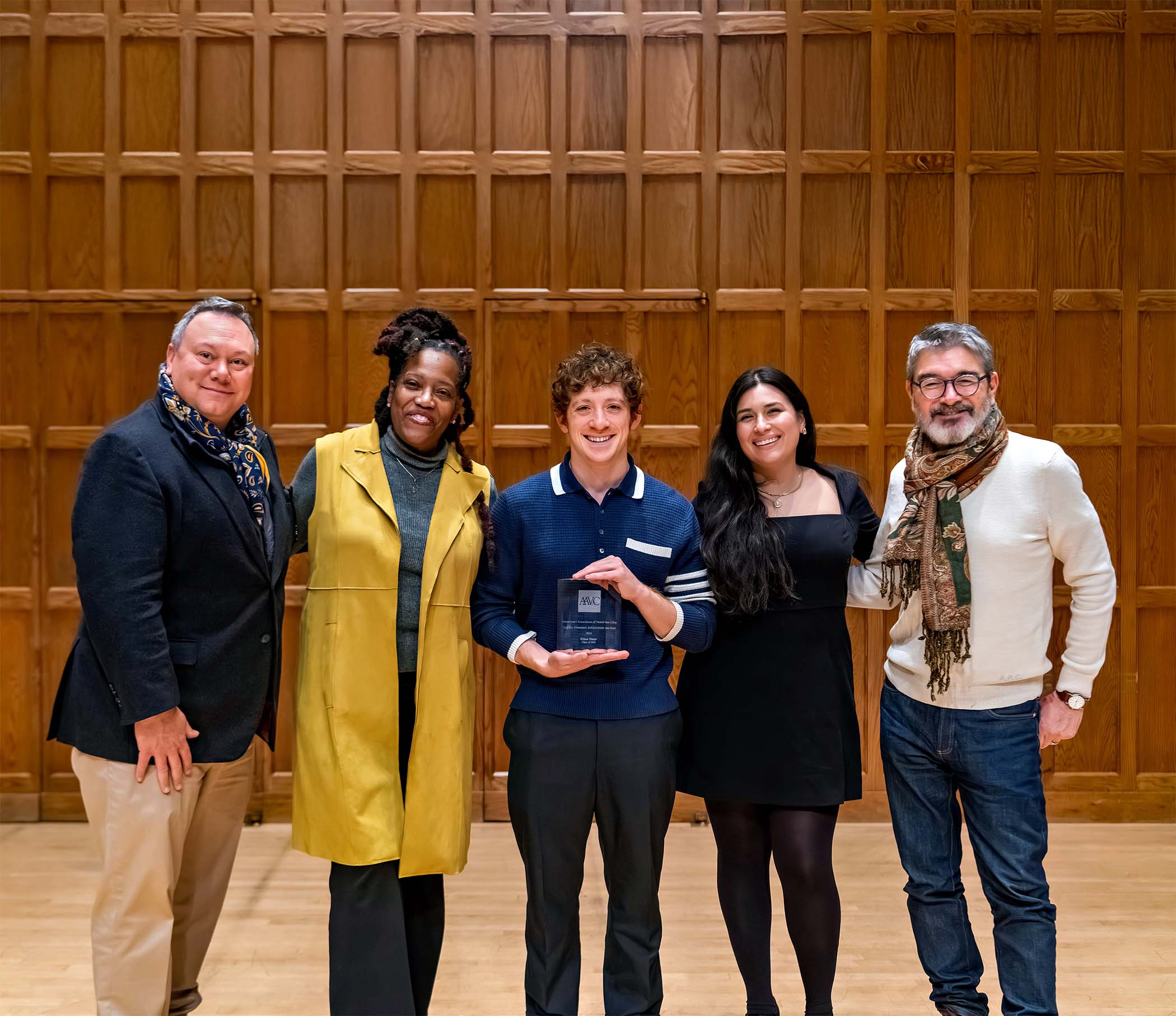 Five people standing together indoors against a wood-paneled wall, smiling, with one person at center holding an award.