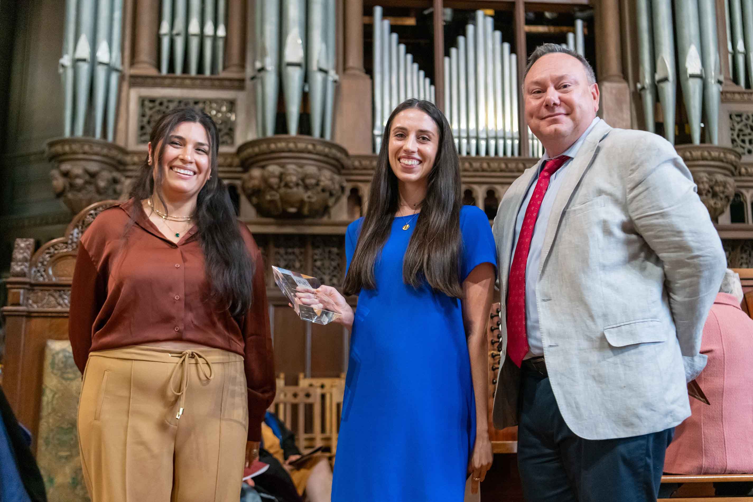 Three people stand in front of a large pipe organ, smiling at the camera, with the person in the middle holding a glass plaque.