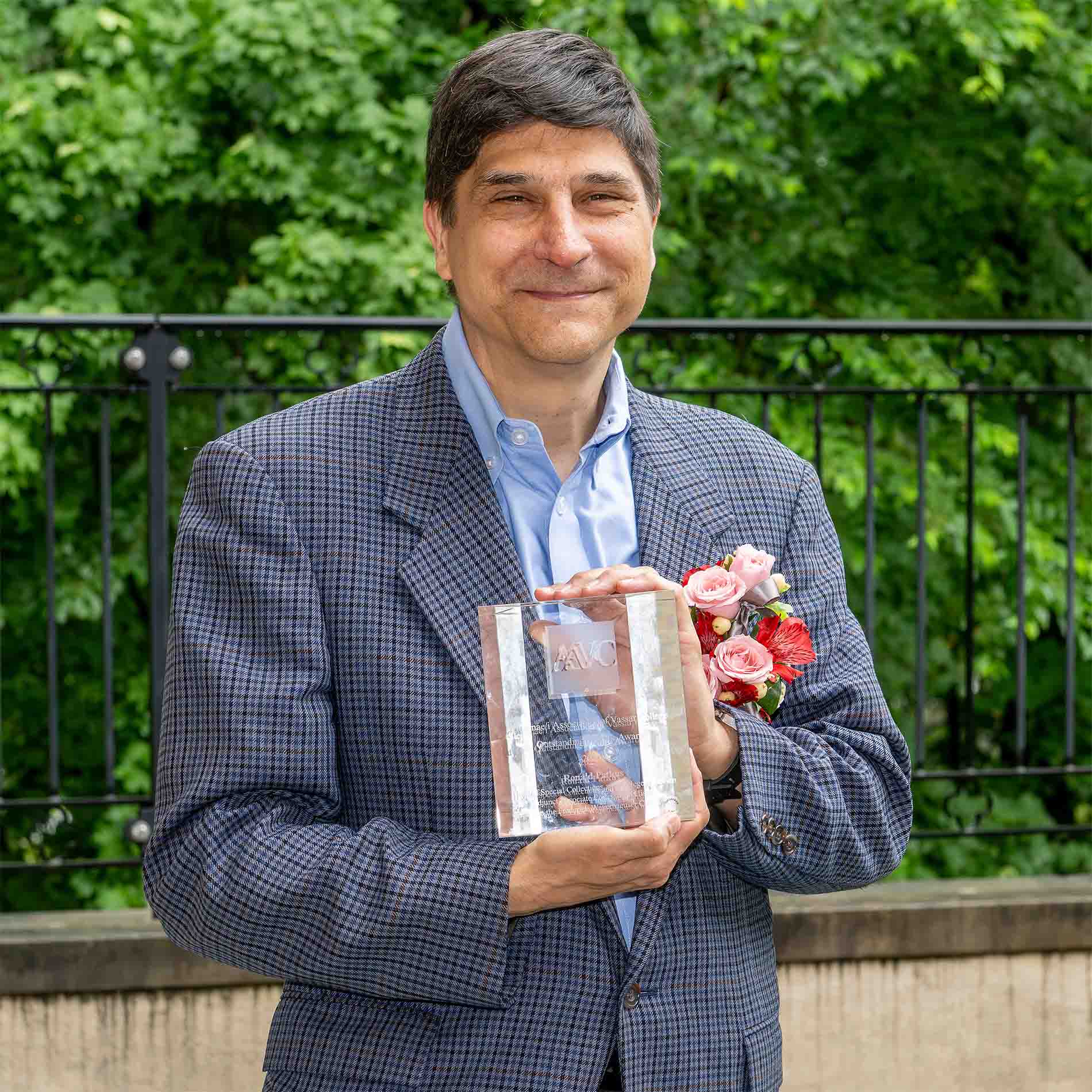 Person standing outdoors holding an award and smiling, with green foliage in the background.