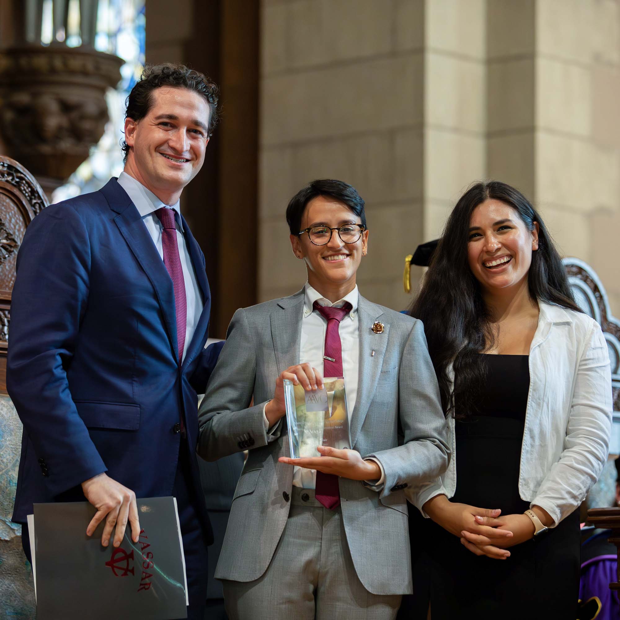 Three people, all with dark brown hair, stand smiling at the camera, with the person in the middle holding a glass plaque.