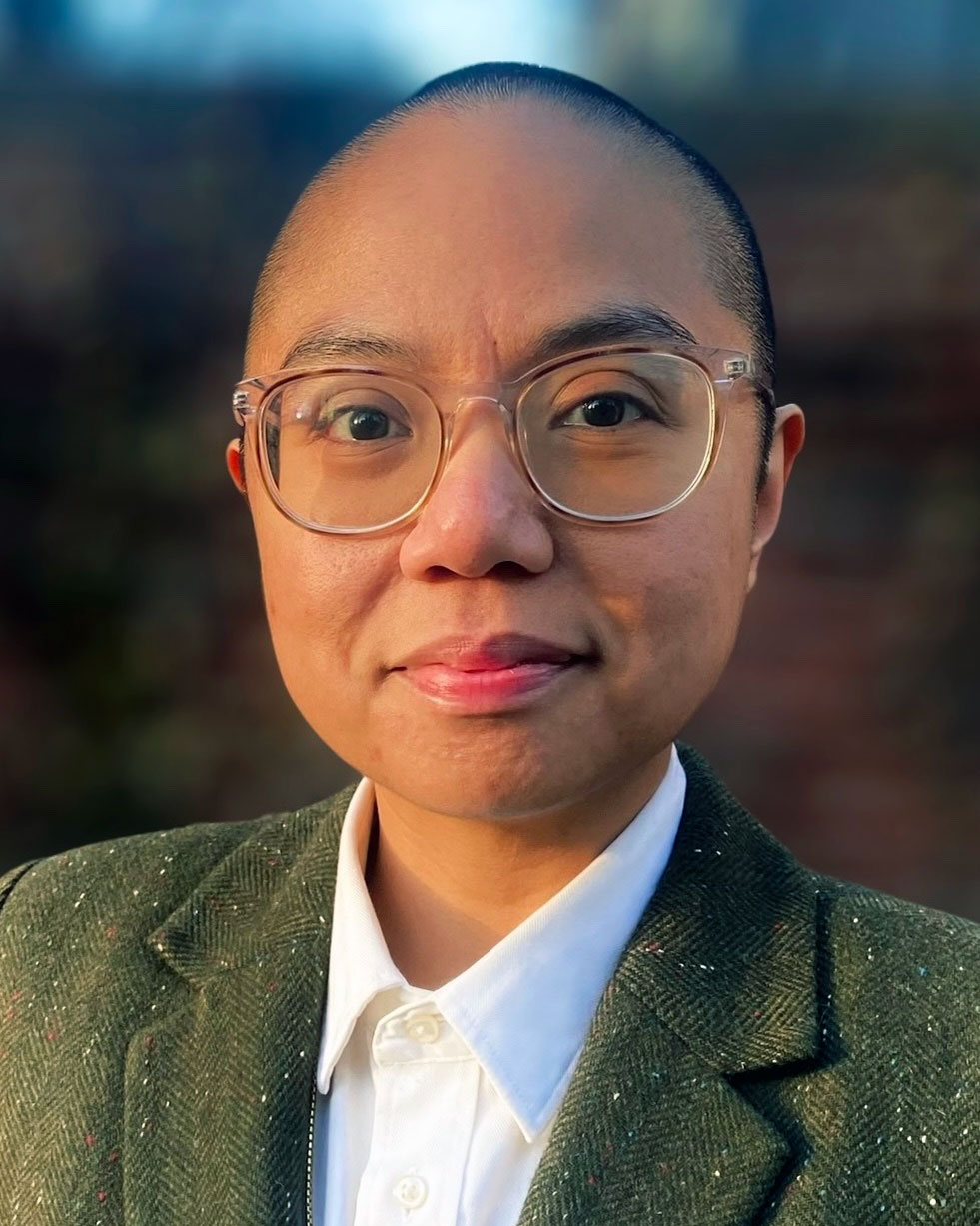 A person with a closely shaven head, glasses, dark colored jacket, and a white shirt smiles at the viewer.