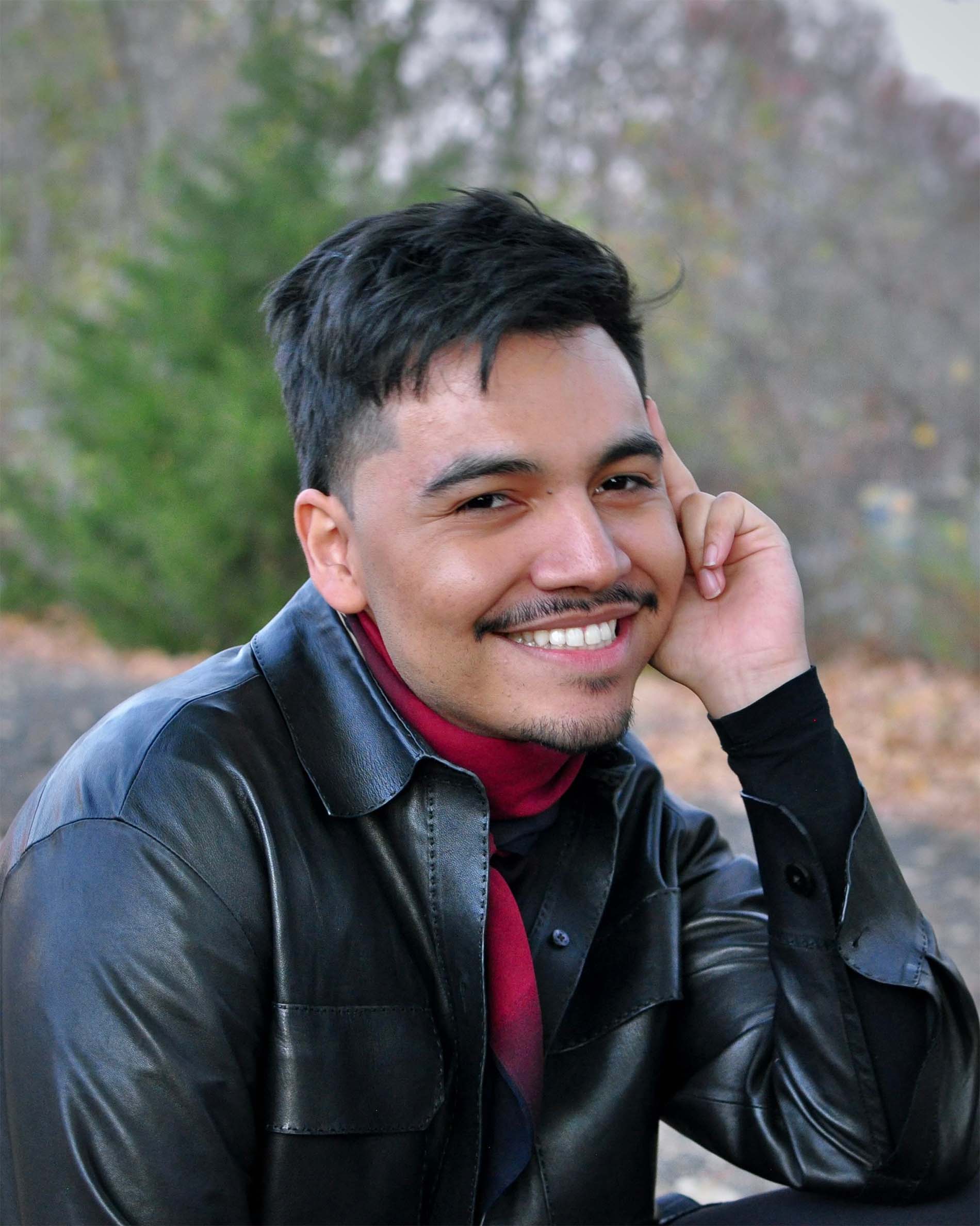 Henry Molina ’19, a person in a black leather jacket and red scarf with short black hair smiling outdoors.