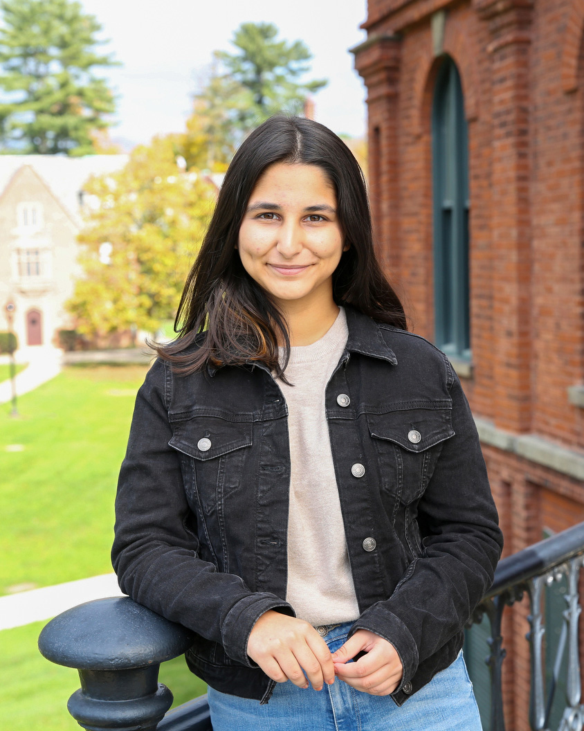 Dhriti Seth ’24, a smiling person with long black hair in a black jacket standing outdoors on stairs of a red brick building.