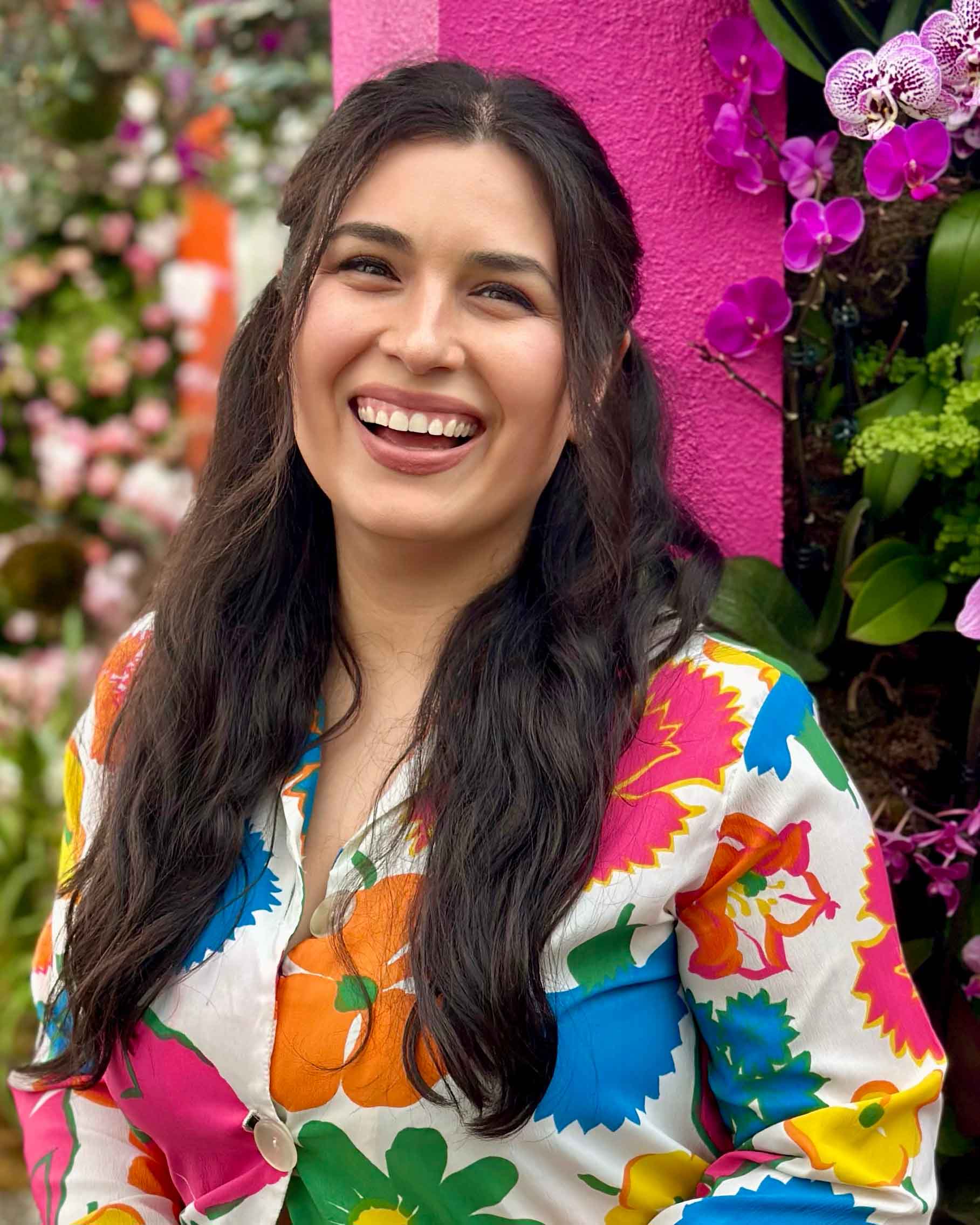 Stephanie Goldberg ’14, a person with long, wavy dark hair smiling in front of a bright pink wall and vibrant orchid flower display, wearing a colorful floral-patterned shirt.