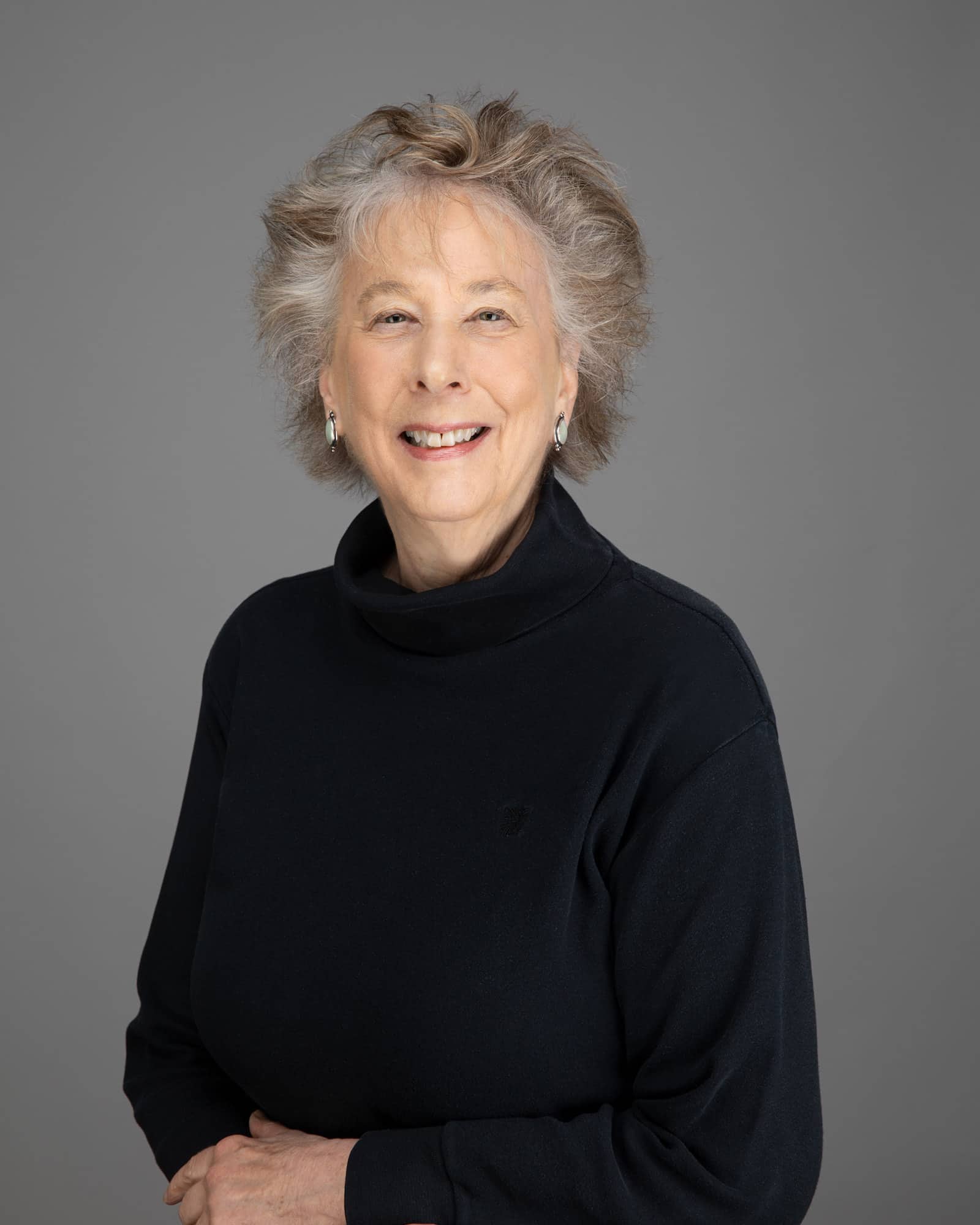 A person with short wavy gray-brown hair and a black turtleneck sweater smiles at the viewer.