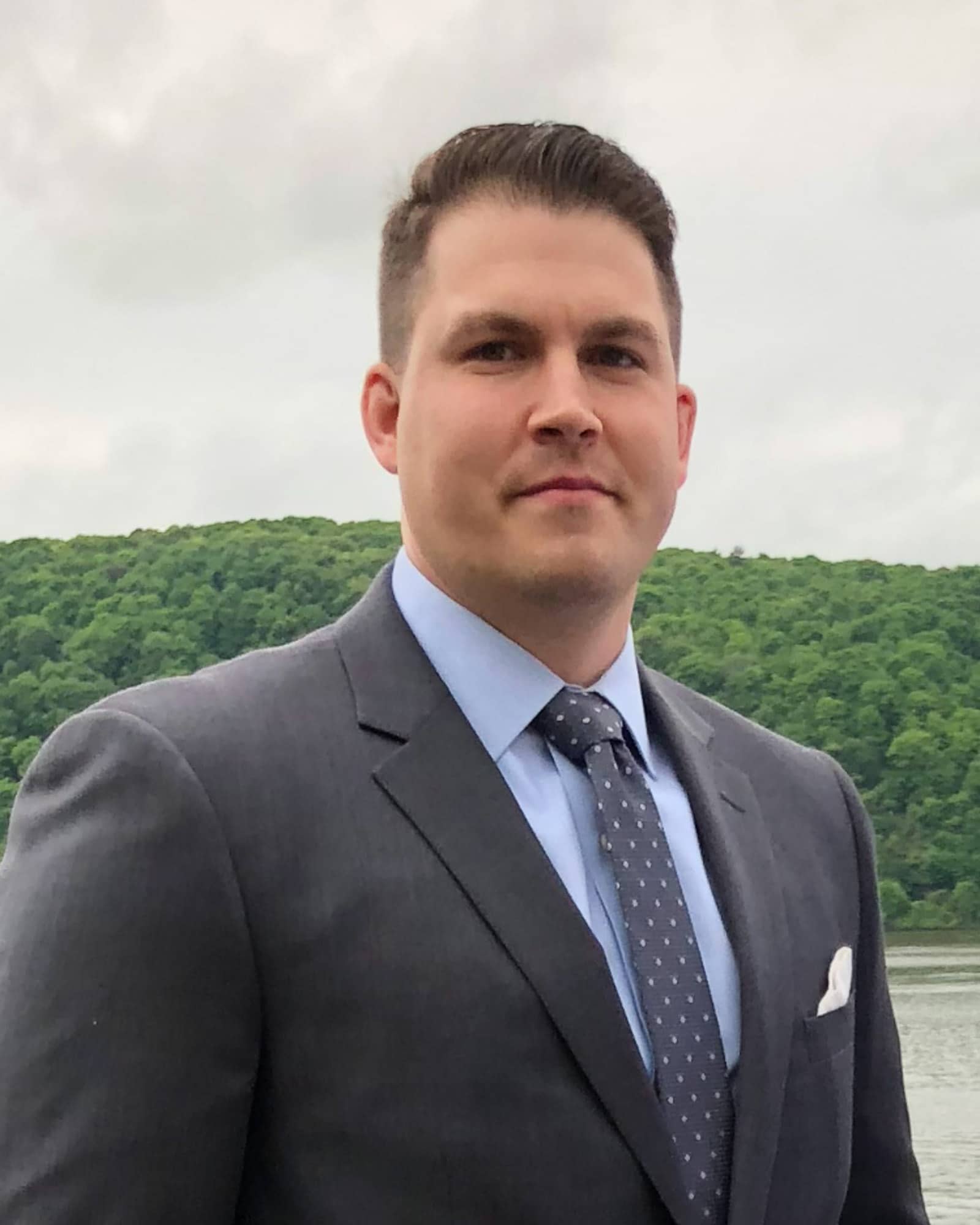 A person wearing a gray suit jacket over a blue shirt and a gray patterned tie looks at the viewer.