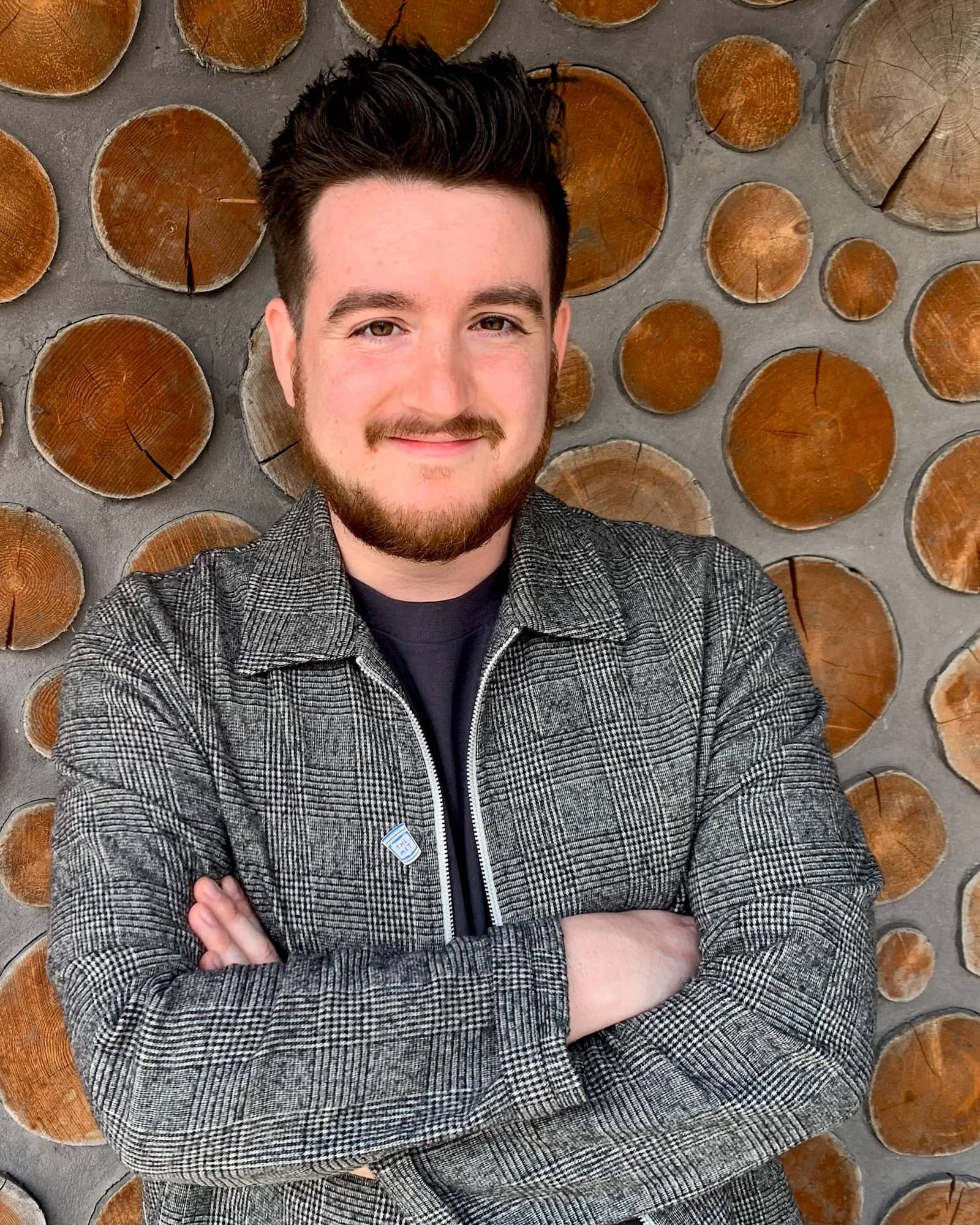 A person with short black hair, a mustache and beard, arms crossed, a gray jacket, and a black shirt smiles at the viewer.