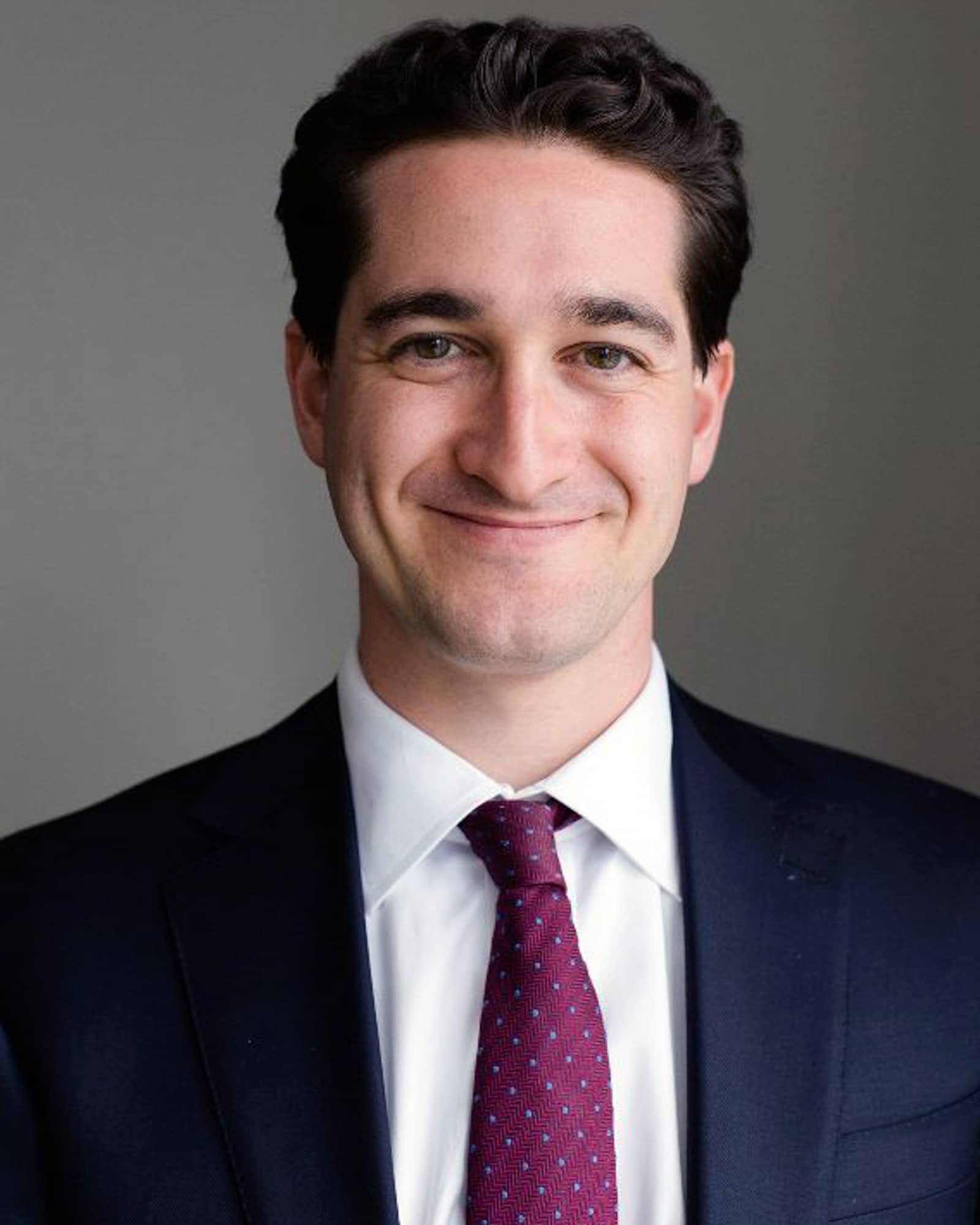 A person with short brown hair, a dark formal jacket, and a burgundy patterned tie smiles at the viewer.