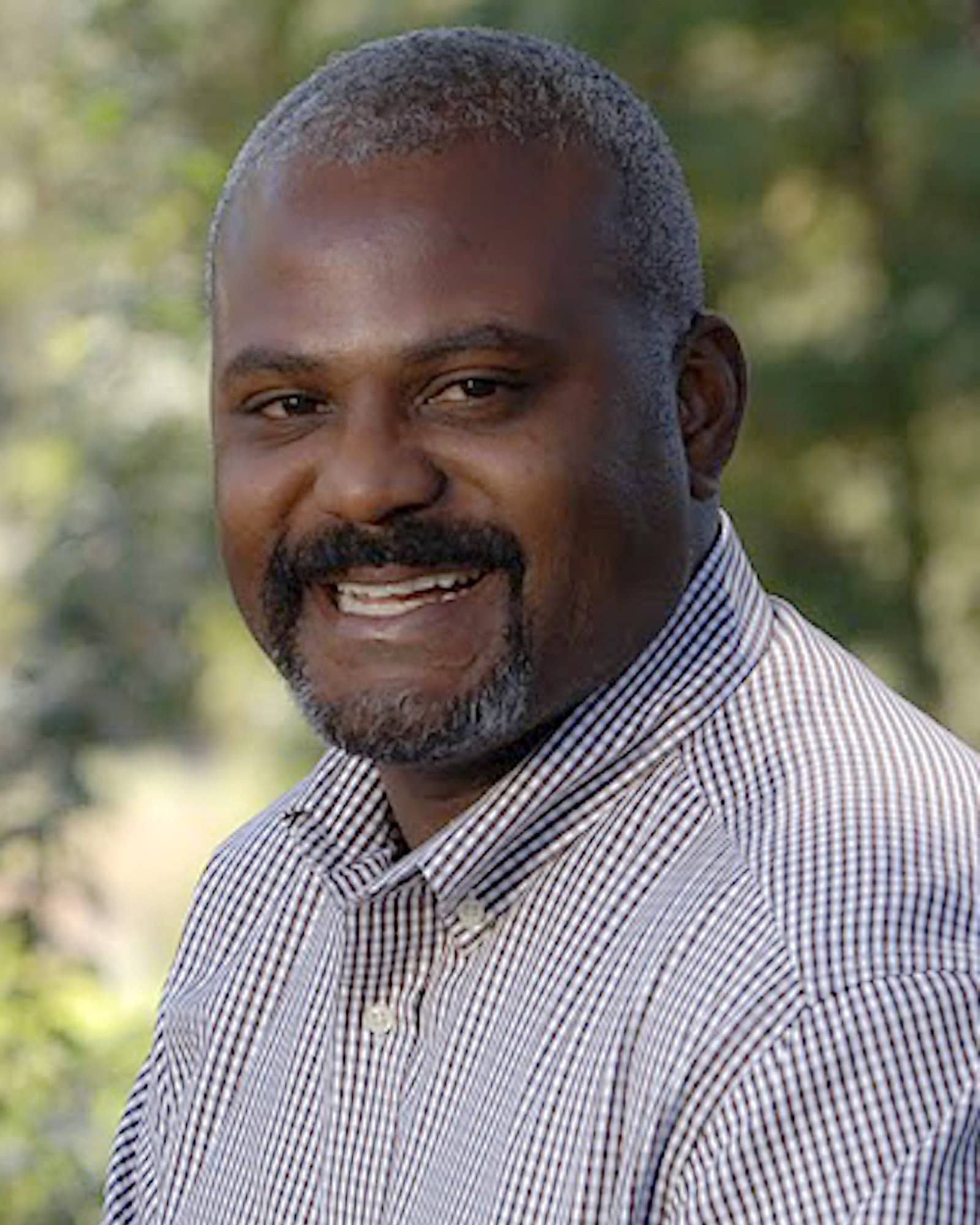 A person with closely cropped dark gray hair, a mustache and goatee, with a black checkered shirt smiles at the viewer.