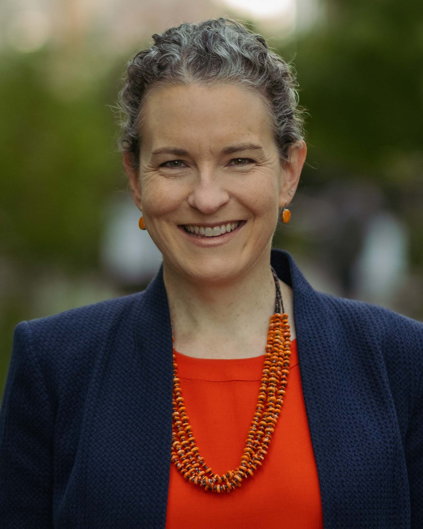 A person with wavy grey-black hair pulled back from their face, a dark blue jacket over a red shirt, and a red beaded necklace smiles at the viewer.