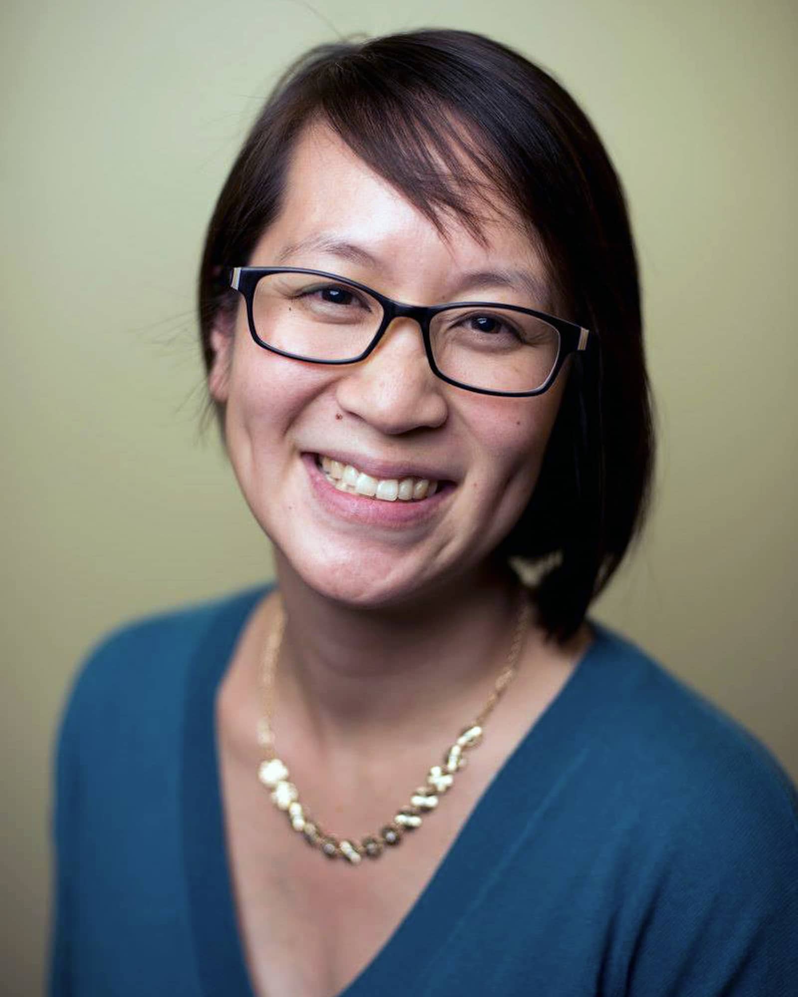 A person with short straight black hair, a dark blue shirt, and a silver necklace smiles at the viewer.