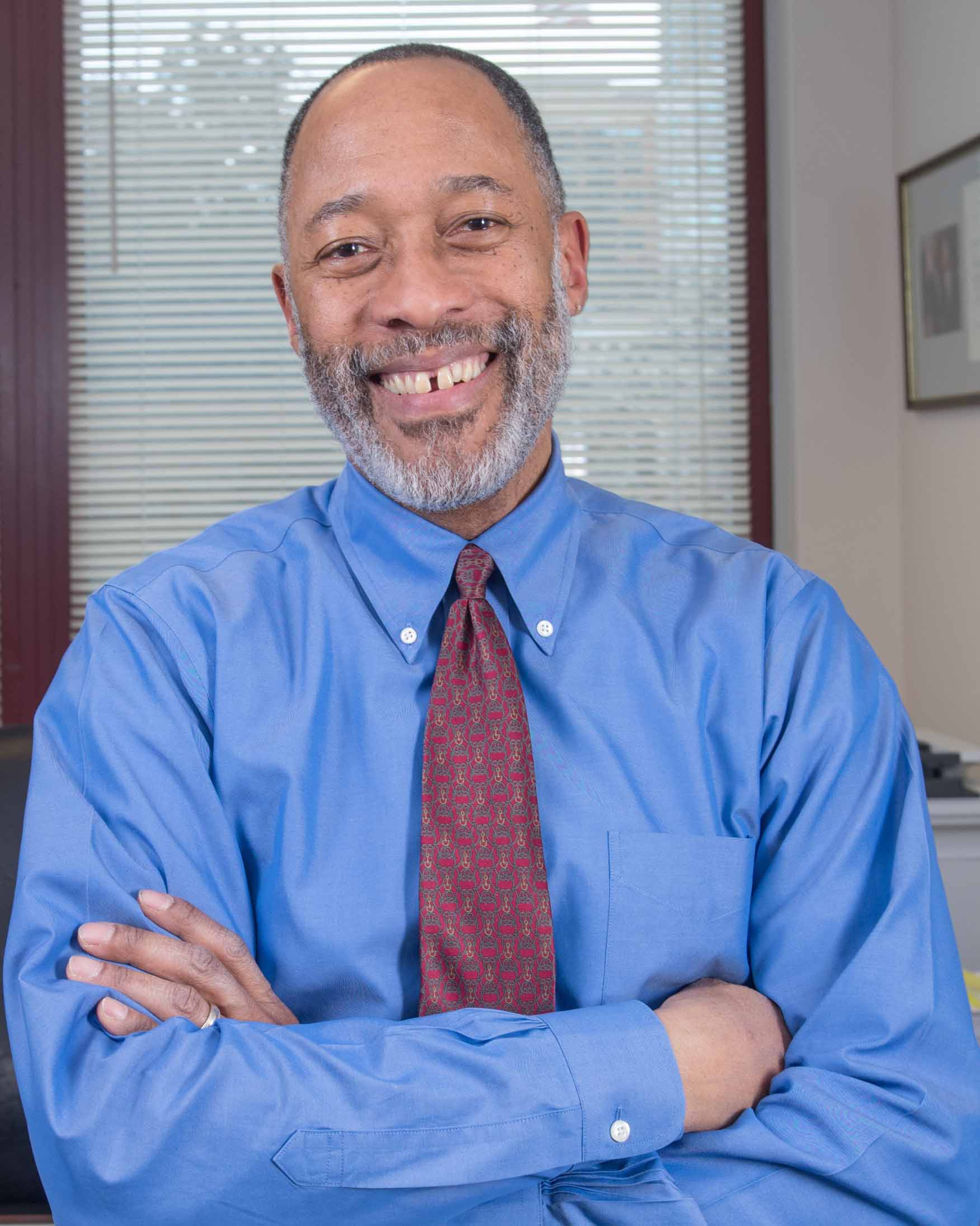 A smiling person with closely cropped gray-black hair, a white beard and gray mustache, wearing a blue shirt with a red, yellow, and gray patterned tie.