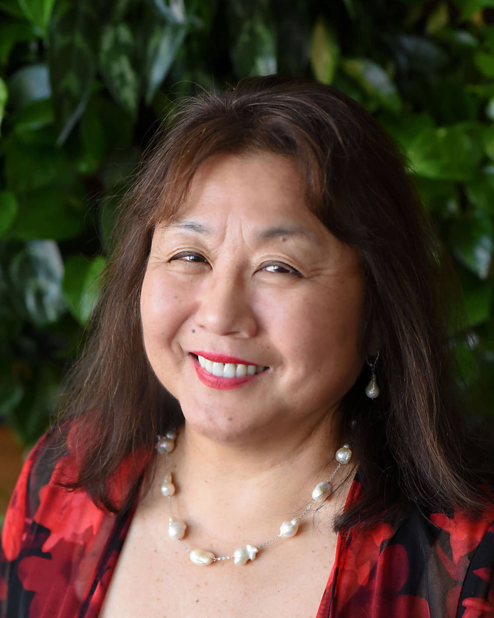 A person with long brown hair and bangs, a red and black shirt, and white bead necklace smiles at the viewer.