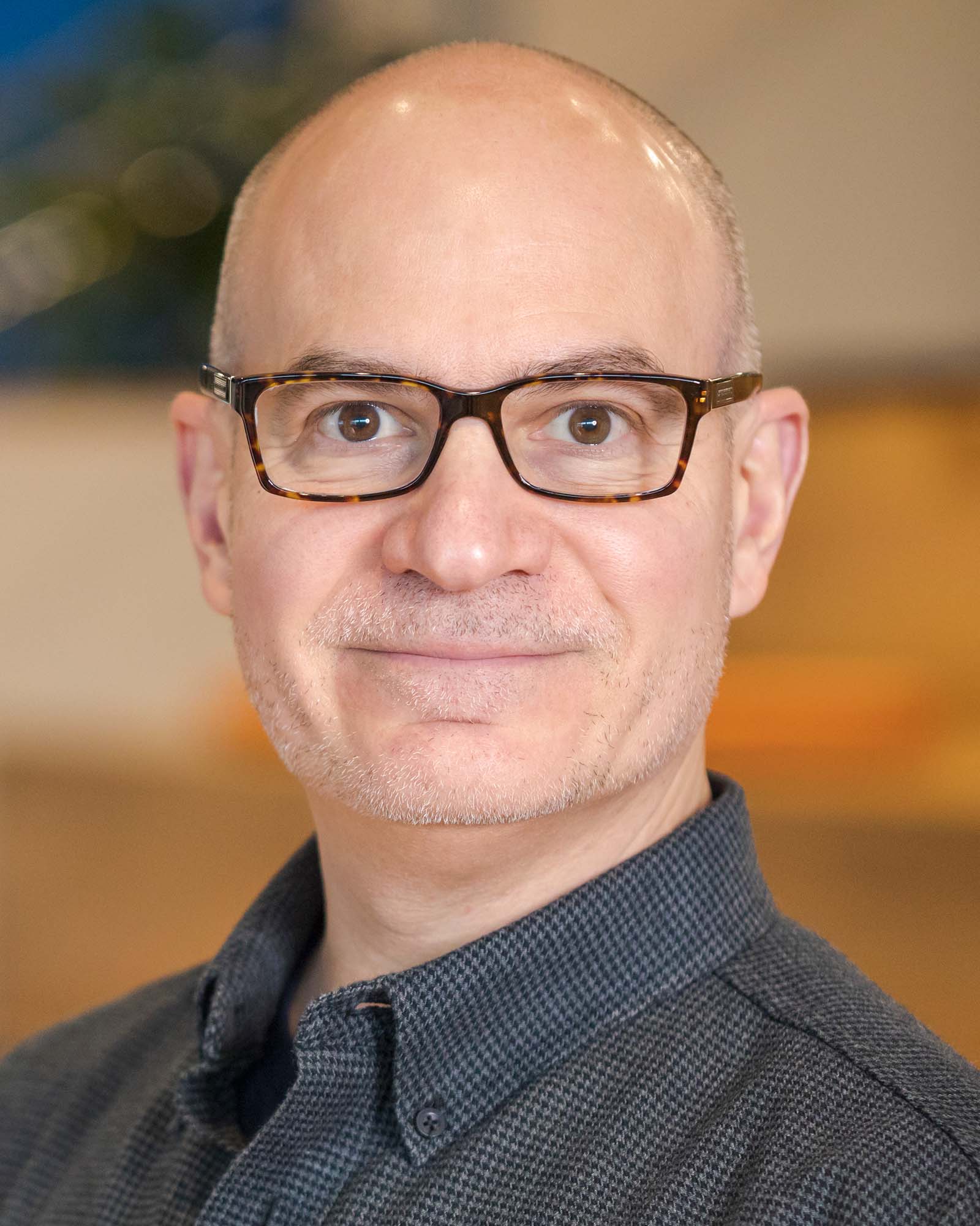 A person with close-shaven hair, beard, and mustache, wearing glasses, smiles at the viewer.