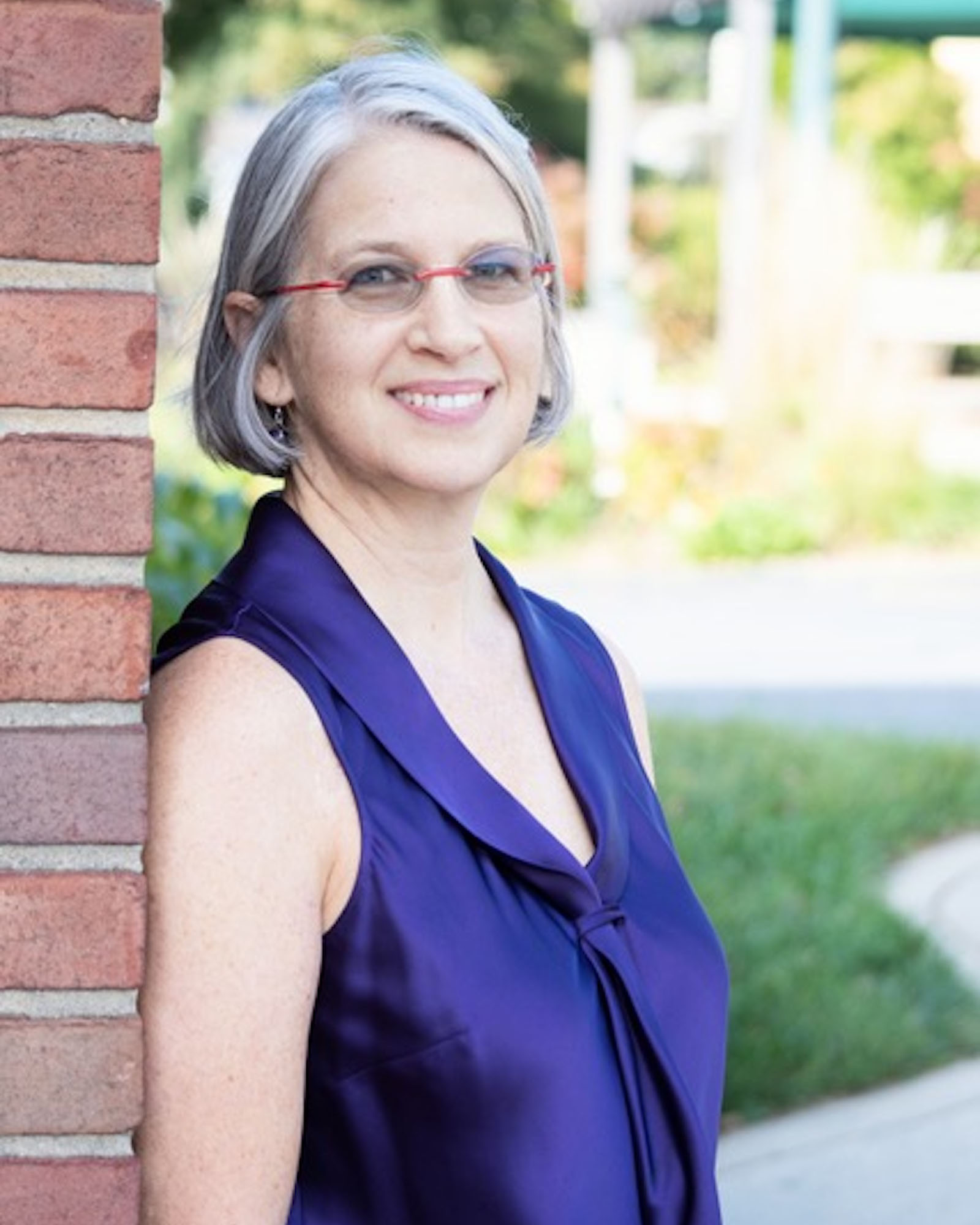 A person with short straight gray and black hair and glasses, wearing a sleeveless dark purple shirt, leans against a brick wall, smiling at the viewer.