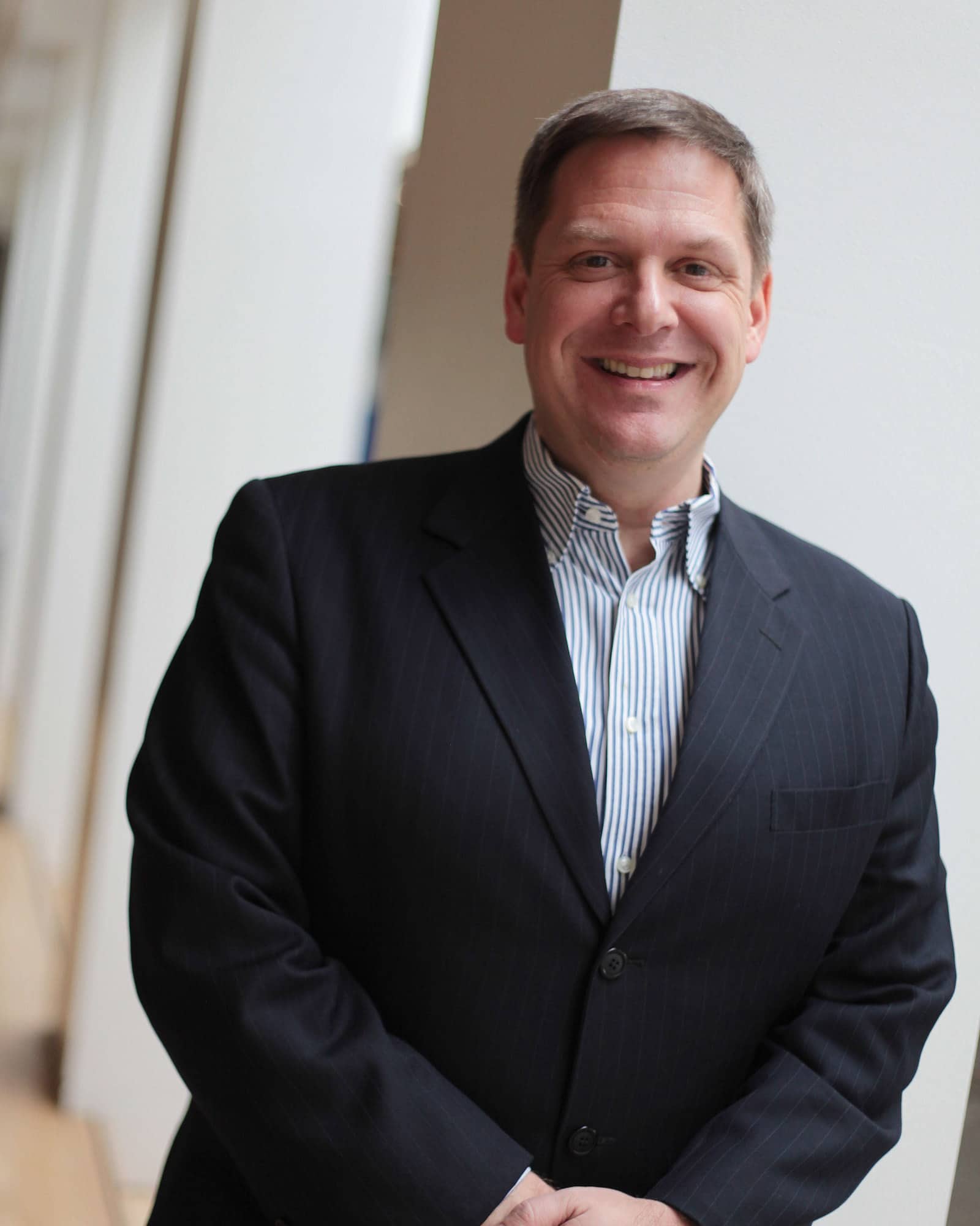 A person with short brown hair, a black jacket, and a blue and white striped shirt smiles at the viewer.