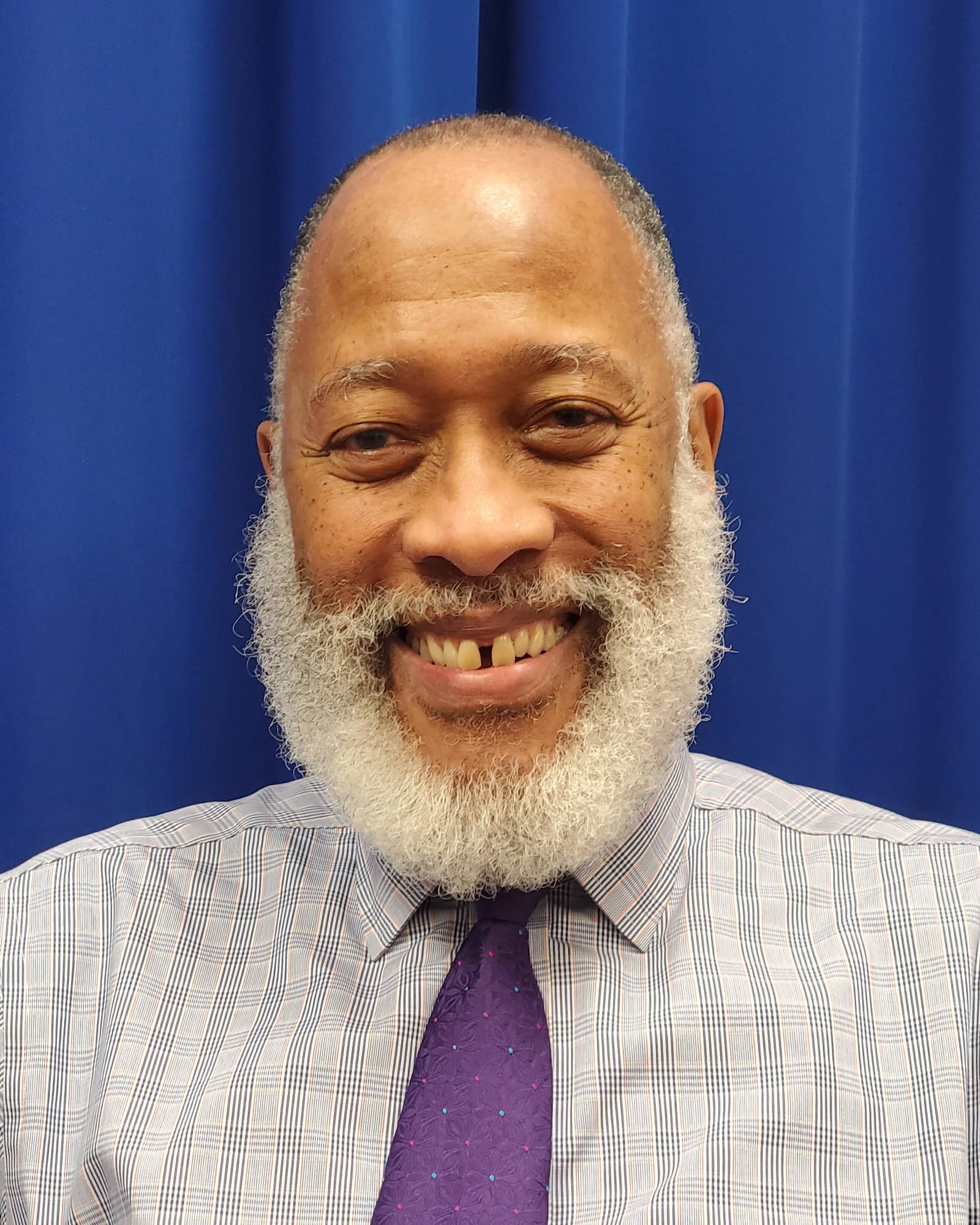 A person with closely cropped gray-black hair, a white beard and gray mustaches, a patterned shirt and purple tie smiles at the viewer.