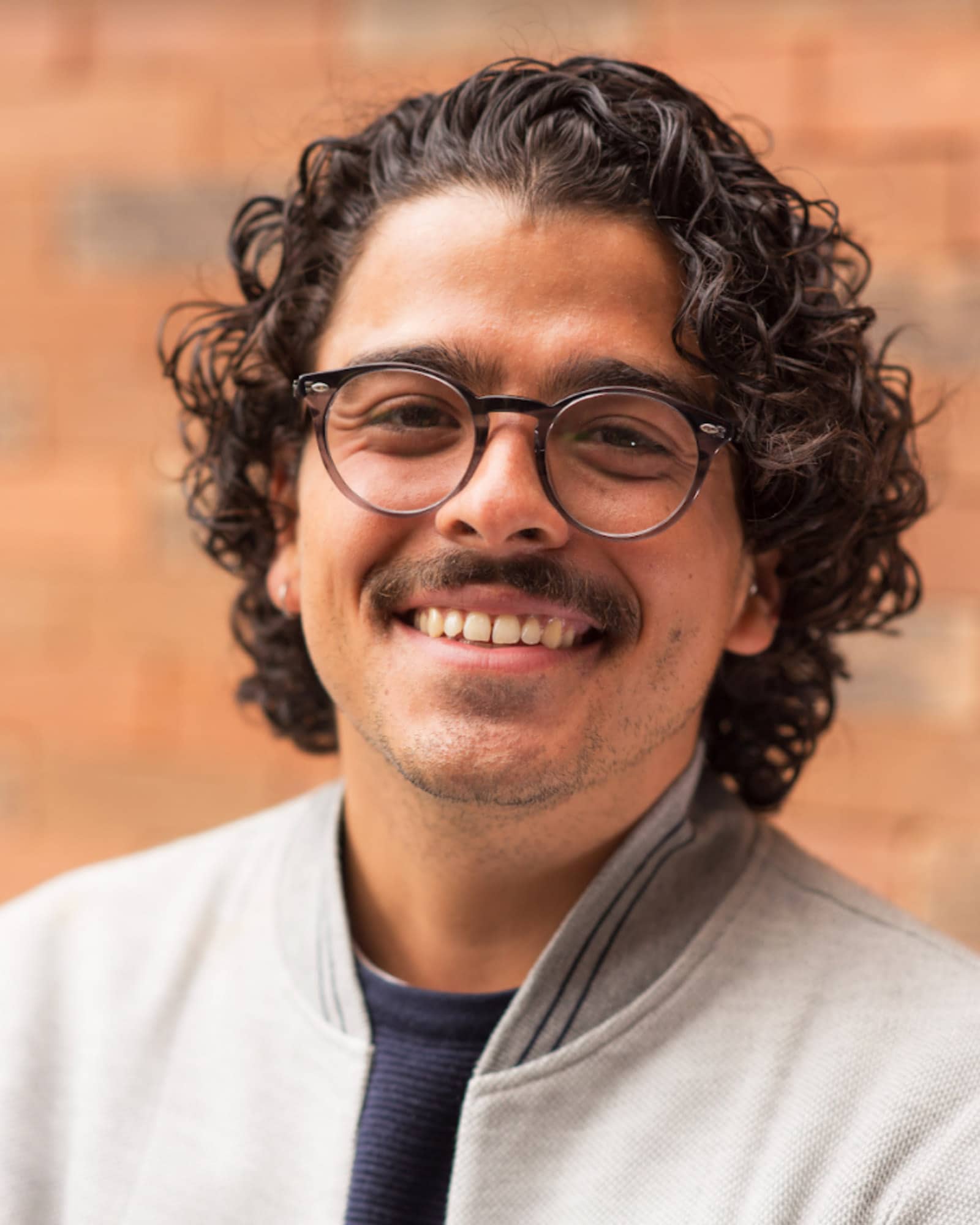 A person with wavy black hair, a mustache, and a light gray jacket over a black shirt smiles at the viewer.