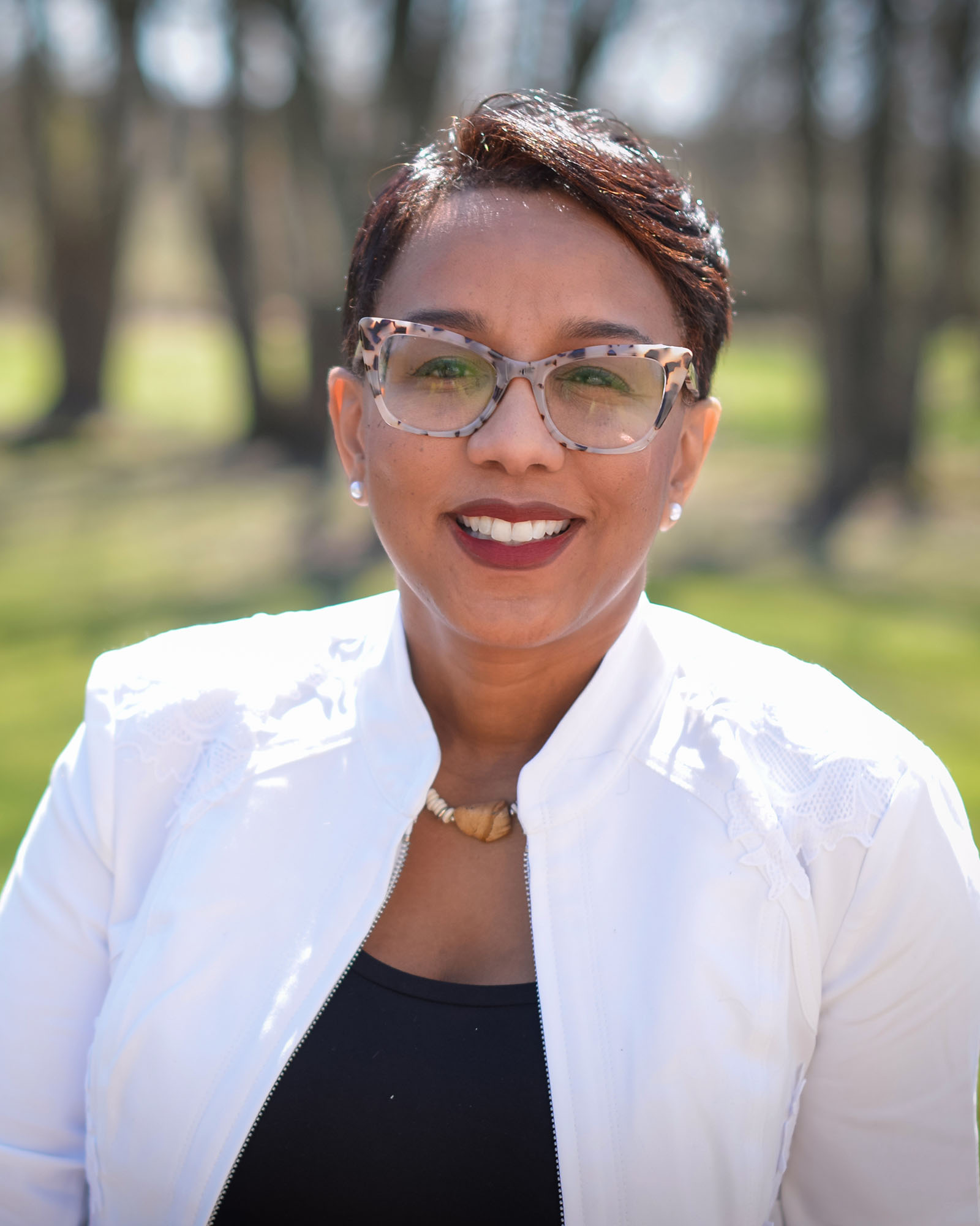 A person with short straight dark hair and glasses wearing a white jacket and black shirt smiles at the viewer.