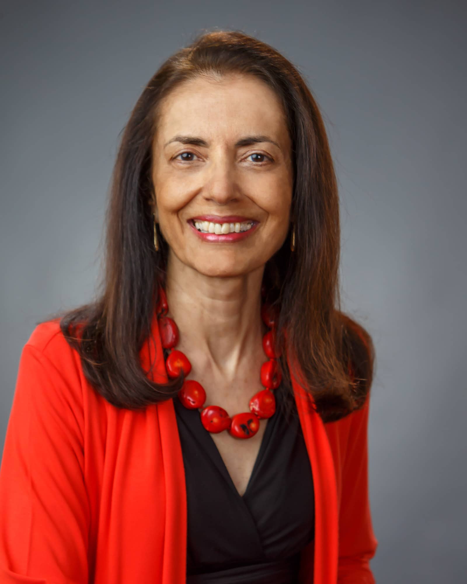 A person with long dark brown hair, a black shirt with a red sweater over it, and a red necklace smiles at the viewer.