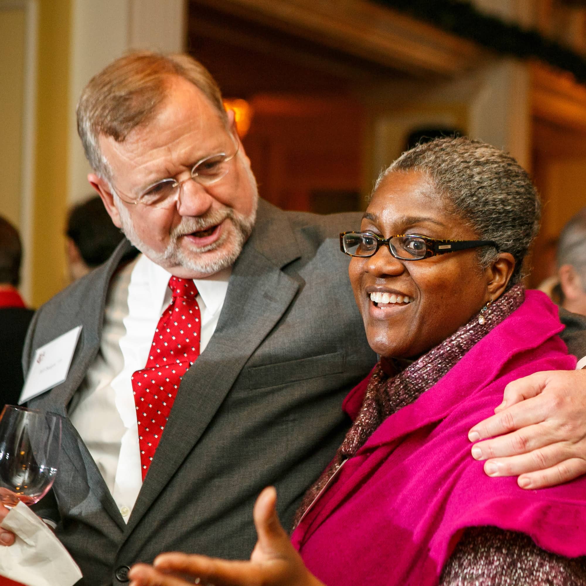 Two people stand with their arms around one another, talking, one with short light brown hair wearing a grey formal jacket and the other with short dark hair and a bright pink jacket.