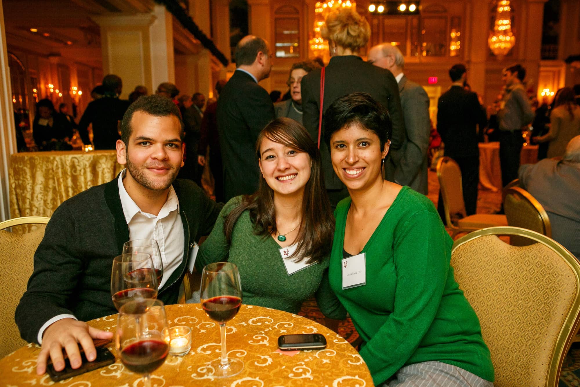 Three people seated at a table with wine glasses on it in a large interior space, smiling and leaning in for a photo.