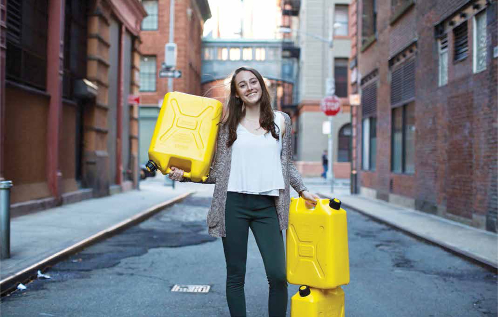 A person with long brown hair, wearing a white shirt, beige sweater, and jean, Sierra Tobin, stands holding bright yellow water jugs in a city alley, smiling at the viewer.
