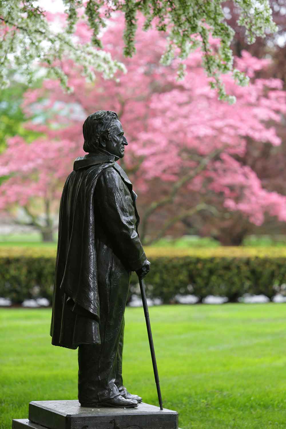 A black metal statue of Matthew Vassar stands in a grassy area with pink trees, visible in the background. The statue is in profile, wearing a cloak and carrying a cane.